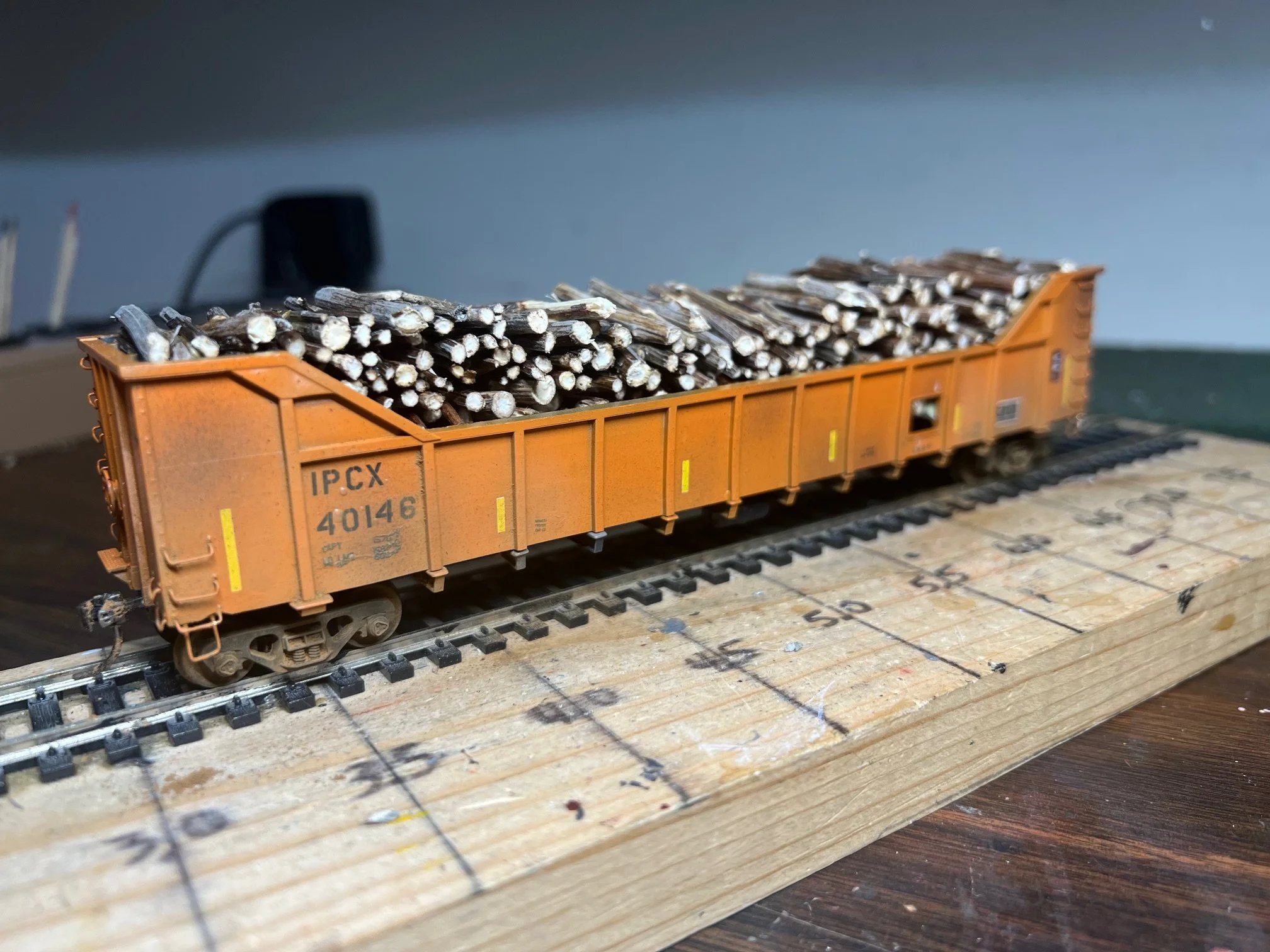 Model train car filled with small wooden sticks, placed on a model railway track, with a measuring ruler on a wooden surface underneath.