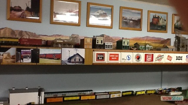 Model train display with framed landscape photographs above, model buildings and train cars, and tracks on multiple levels.