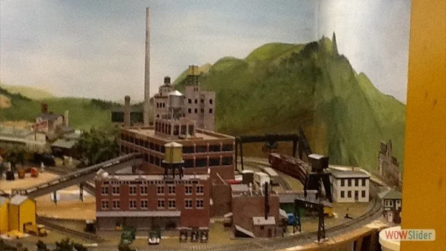 Model train setup with a factory, train tracks, and painted green hills in the background.