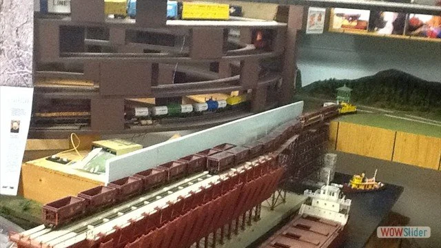 Model train setup with tracks, cars, and a small boat in a miniature landscape.