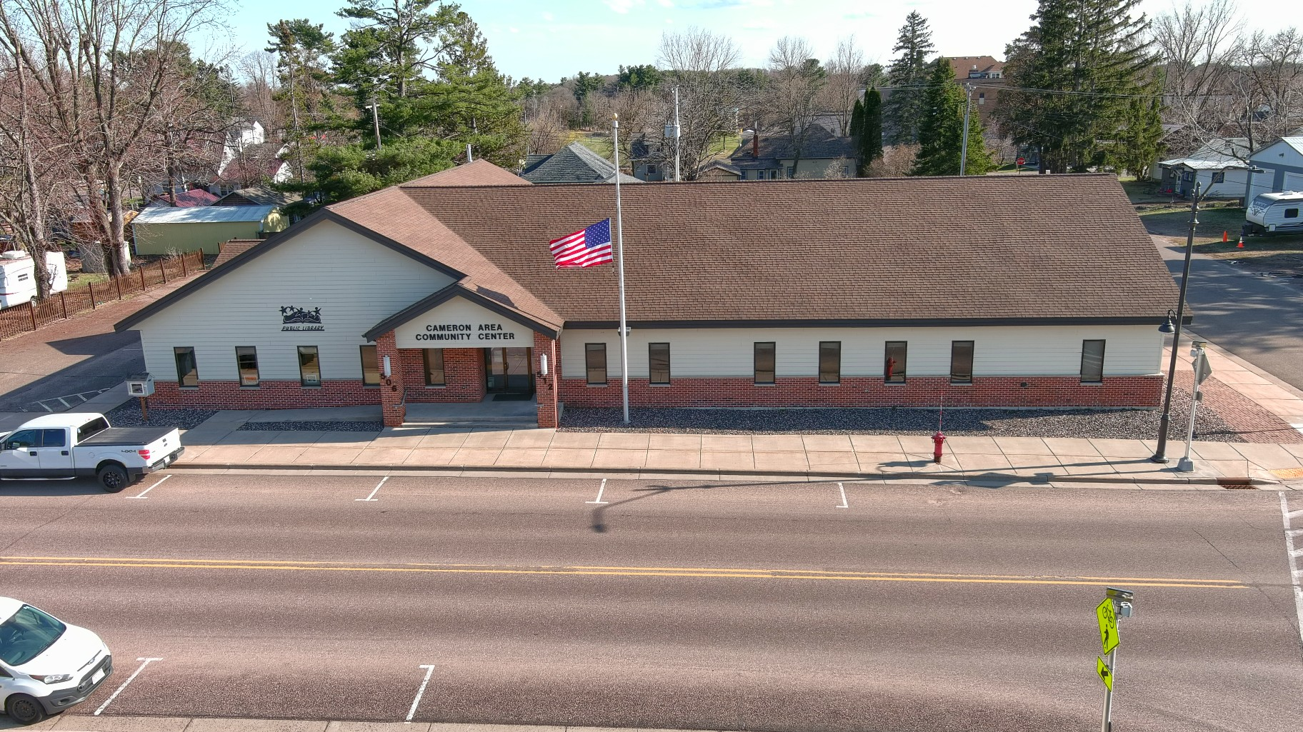 An aerial view of the Cameron Area Community Center, a one-story building with a brick and siding exterior, a brown shingled roof, and an American flag flying in front. The building has a sign that reads "Cameron Area Community Center" and a smaller sign for "Public Library." A sidewalk runs along the front, with a small parking lot and a fire hydrant. There are trees and other buildings in the background, and the street in front has parked cars and bicycle lane signage.