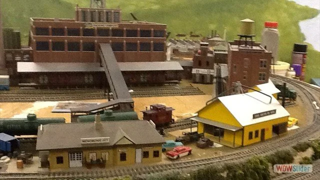 Model train layout with a yellow train station, train tracks, a yellow building, and a large industrial building in the background.