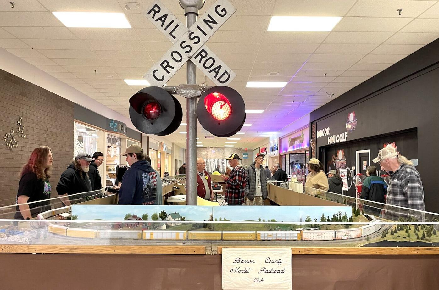 Indoor model train exhibit with a railway crossing signal, people viewing the train setup, and shops in the background.