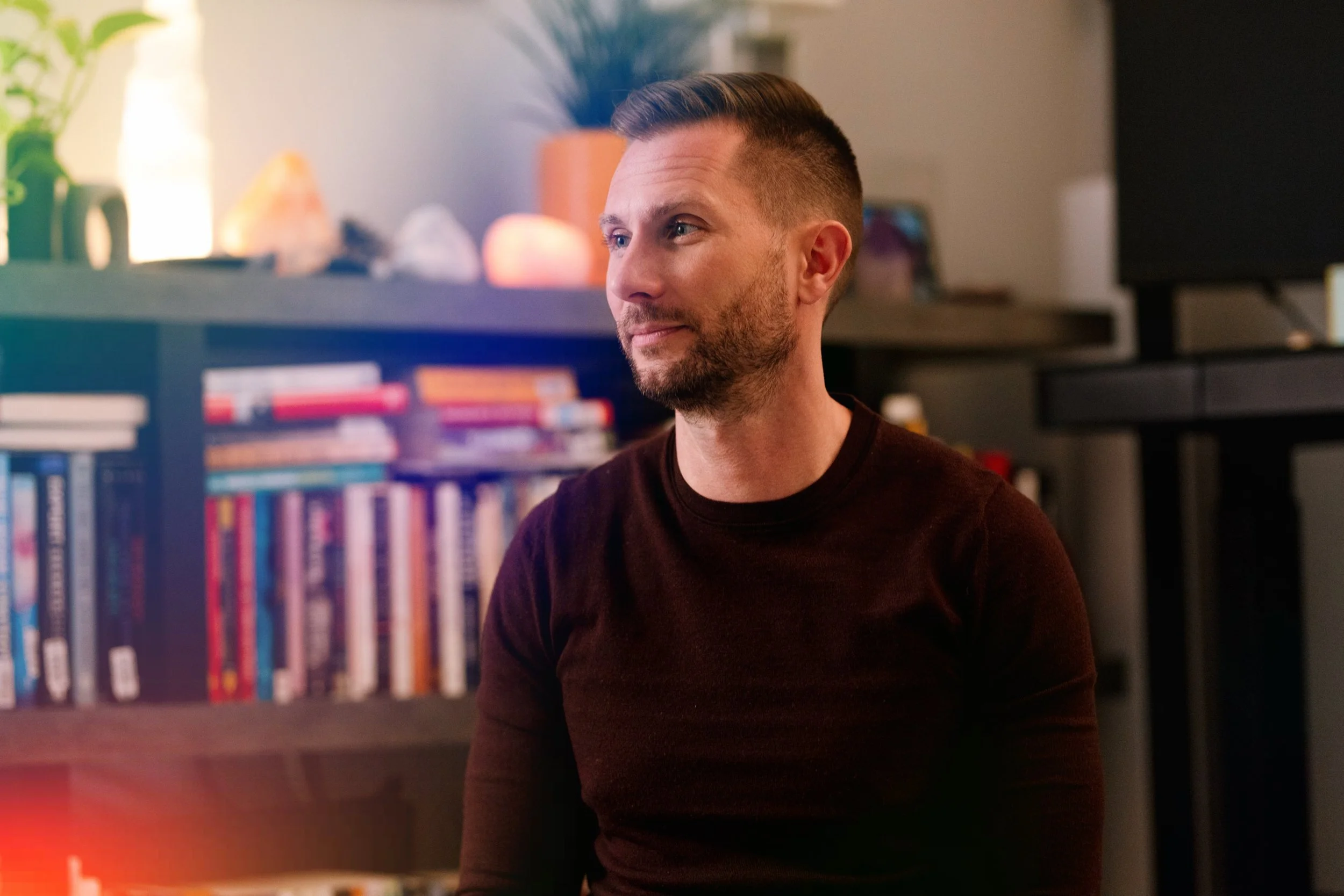 A man with short dark hair and a beard, wearing a dark shirt, sits in front of a bookshelf with colorful books and a potted plant in the background.