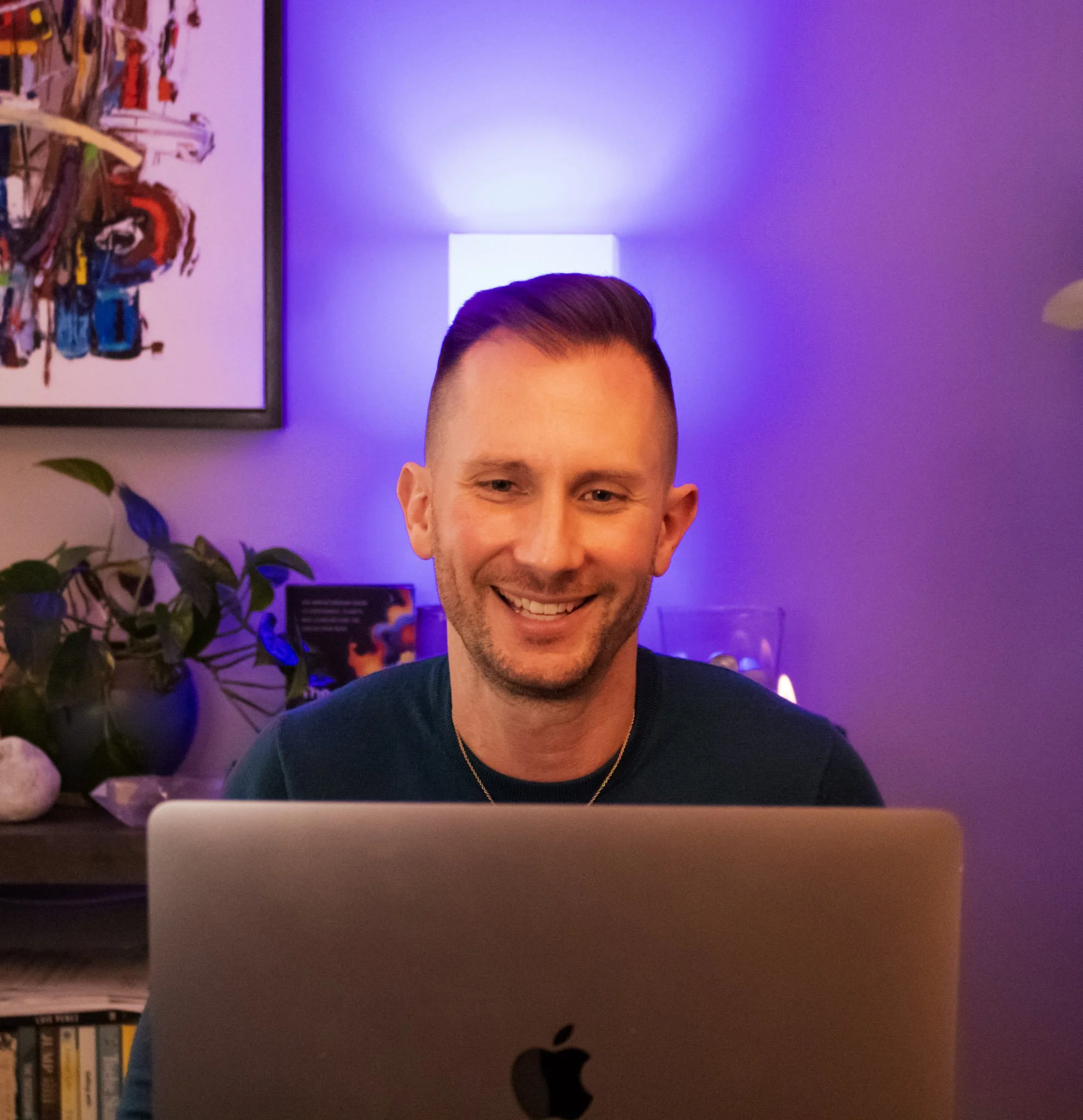 Partially visible person with slicked back hair smiling, seen over a silver Apple MacBook laptop on a desk with a black Apple logo, surrounded by a bookshelf, a purple wall with a square light and a purple light.