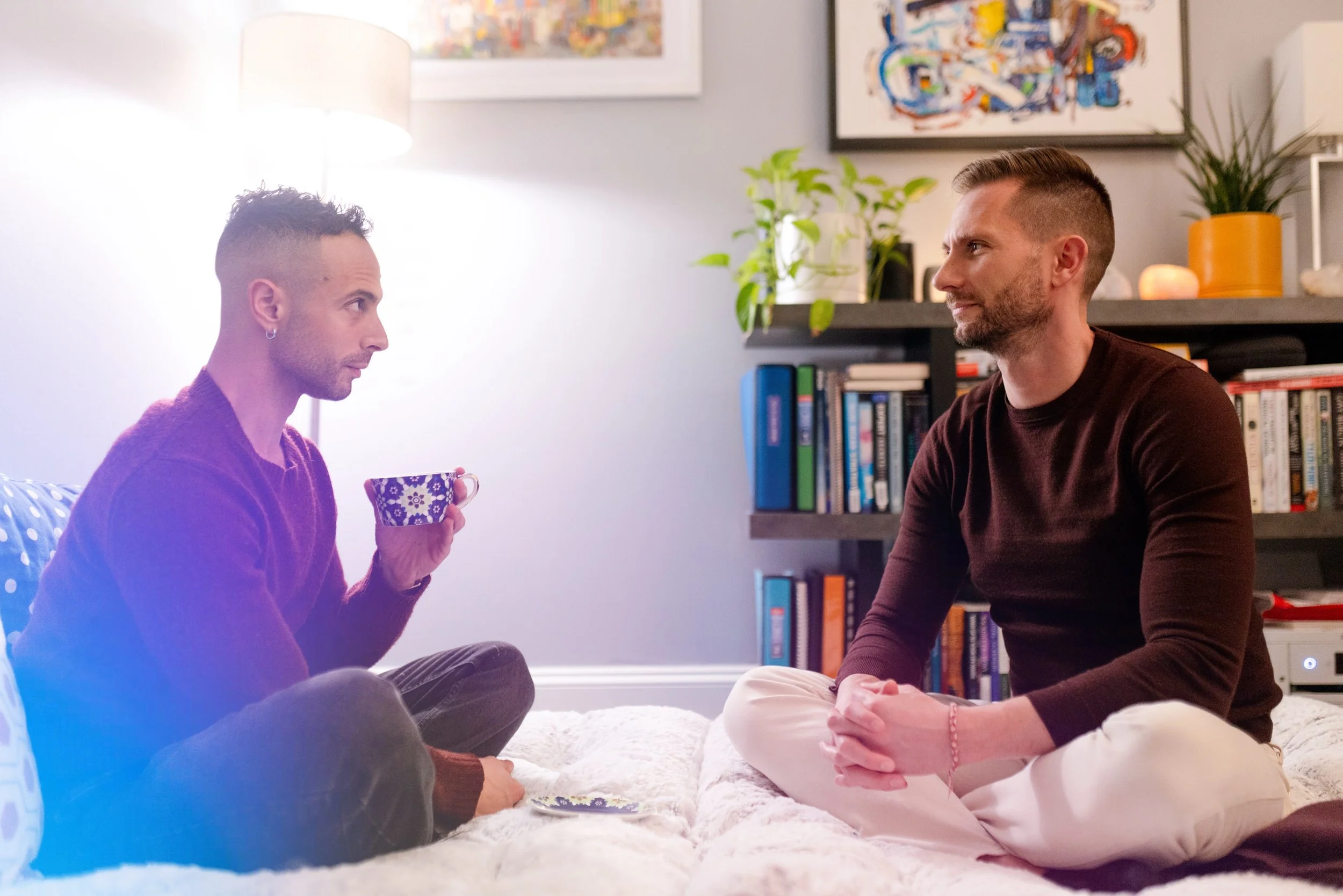 Two men sit on a white carpet in a living room, facing each other, with potted plants and abstract art on the wall behind them; one man holds a mug.