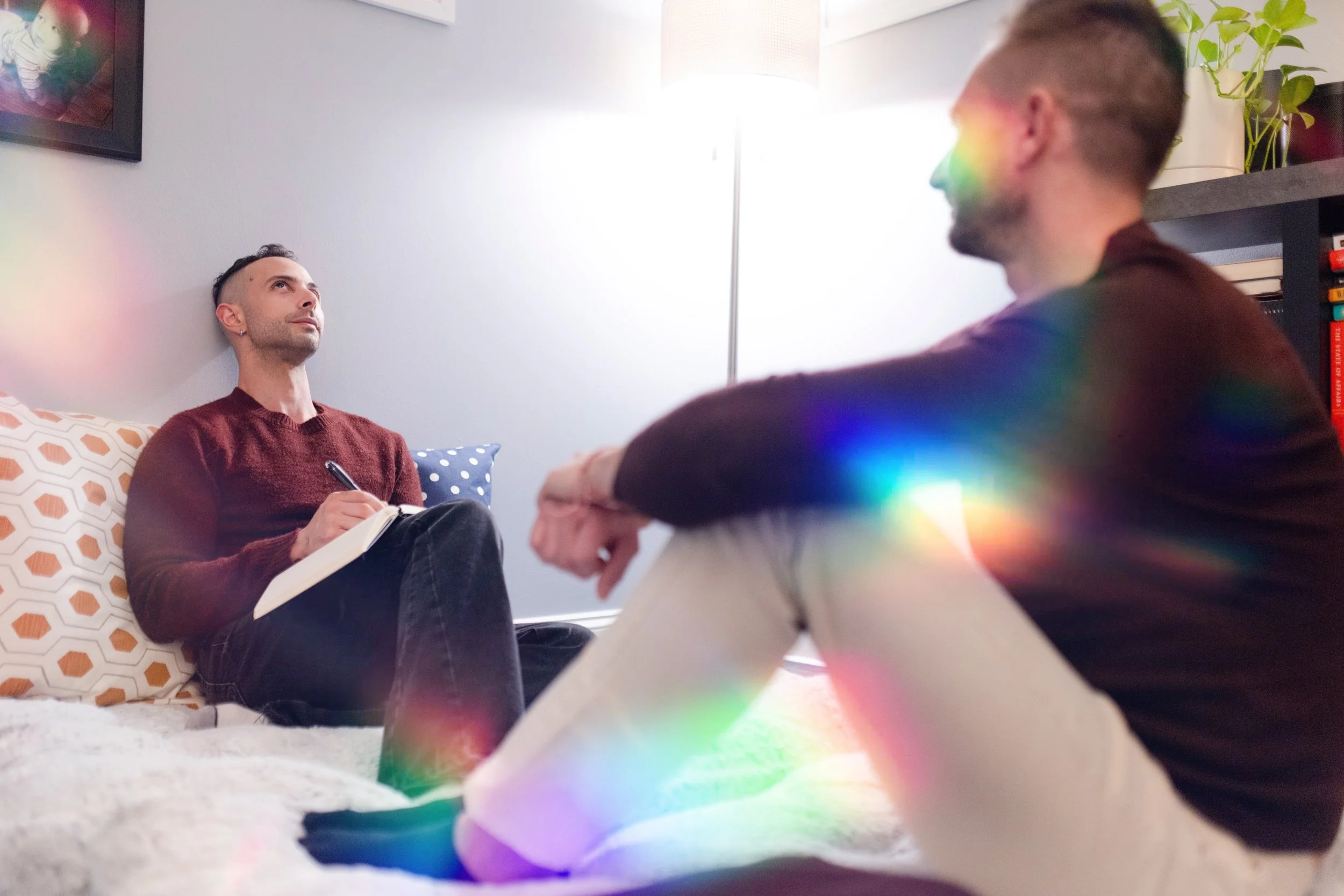 Two young men having a conversation in a cozy living room with a white wall, a framed picture, and a bookshelf. One man is sitting on a sofa with geometric-patterned pillows, holding a notebook and pen, while the other is sitting on the floor with blurred rainbow light effects in the foreground.