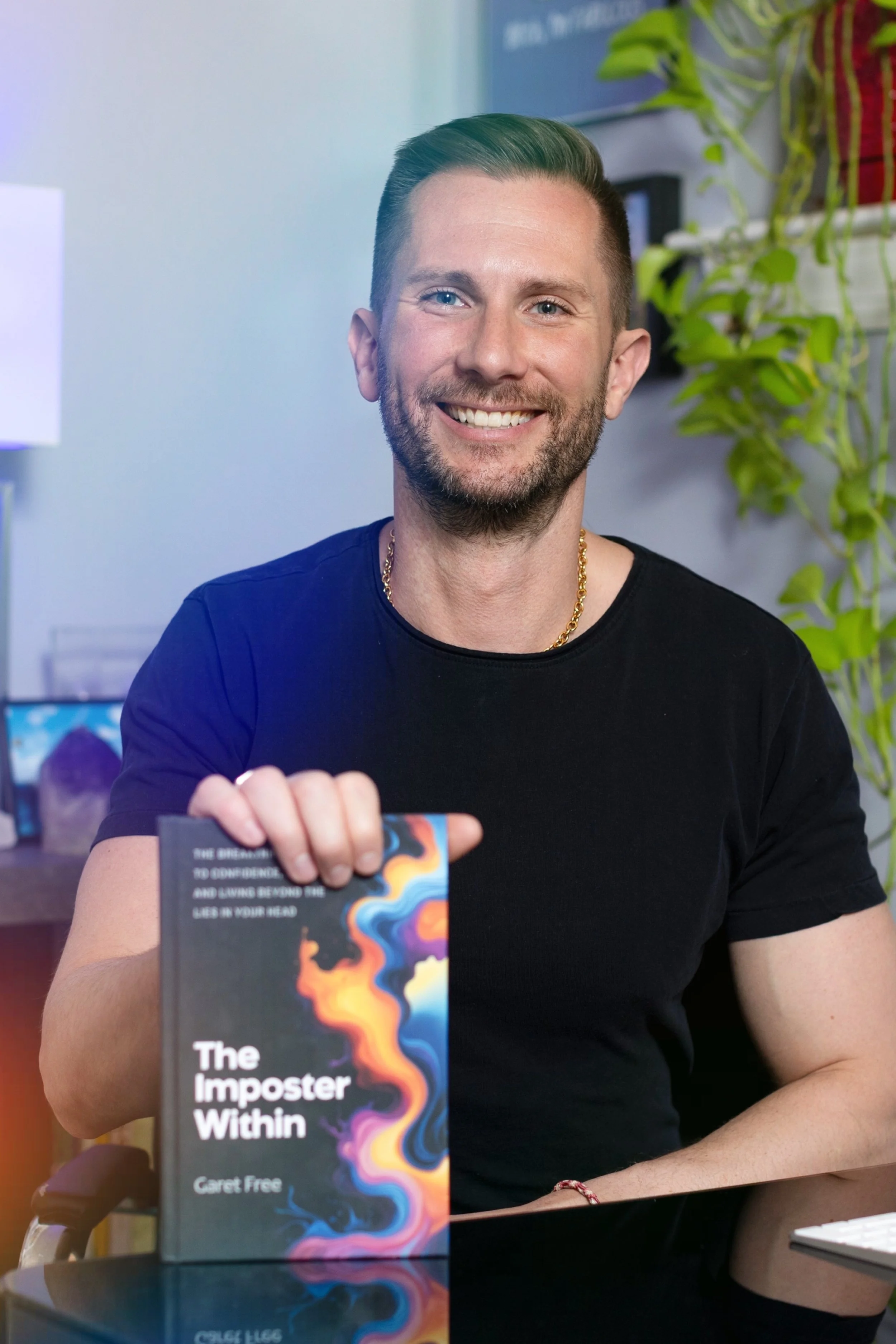 A smiling man with a beard and short hair, wearing a black shirt and gold chain, sitting at a table in an office setting. He is holding a colorful book titled "The Imposter Within" by Garett Free. There are plants and office items in the background.