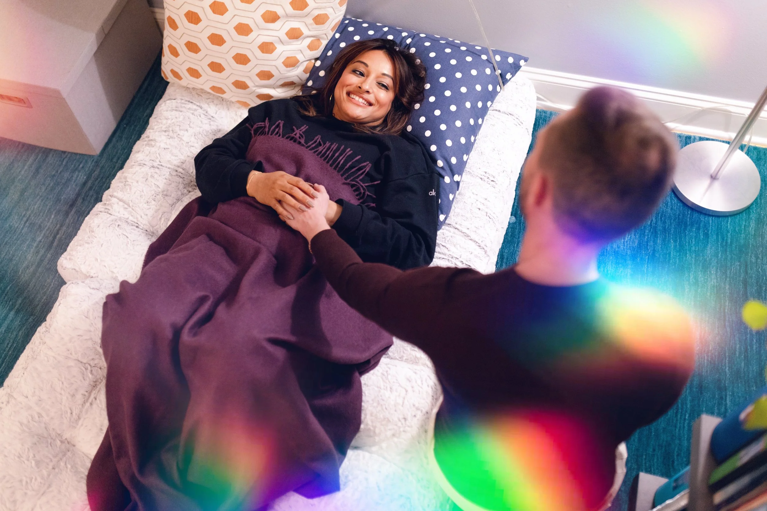 A woman lying on a bed with her head on a blue polka dot pillow, smiling, holding hands with a person whose back is to the camera, in a room with colorful lighting and a white textured blanket.