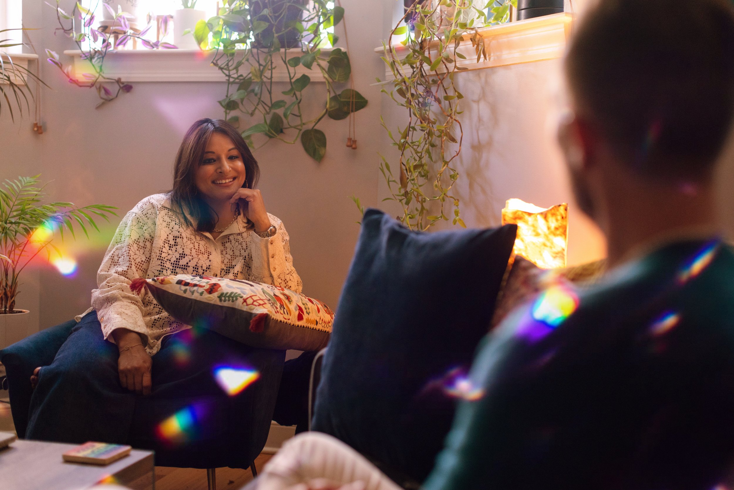 A woman with shoulder-length brown hair smiling, sitting cross-legged on the floor in a cozy room with plants and colorful string lights, engaging in a conversation with another person whose back is visible.
