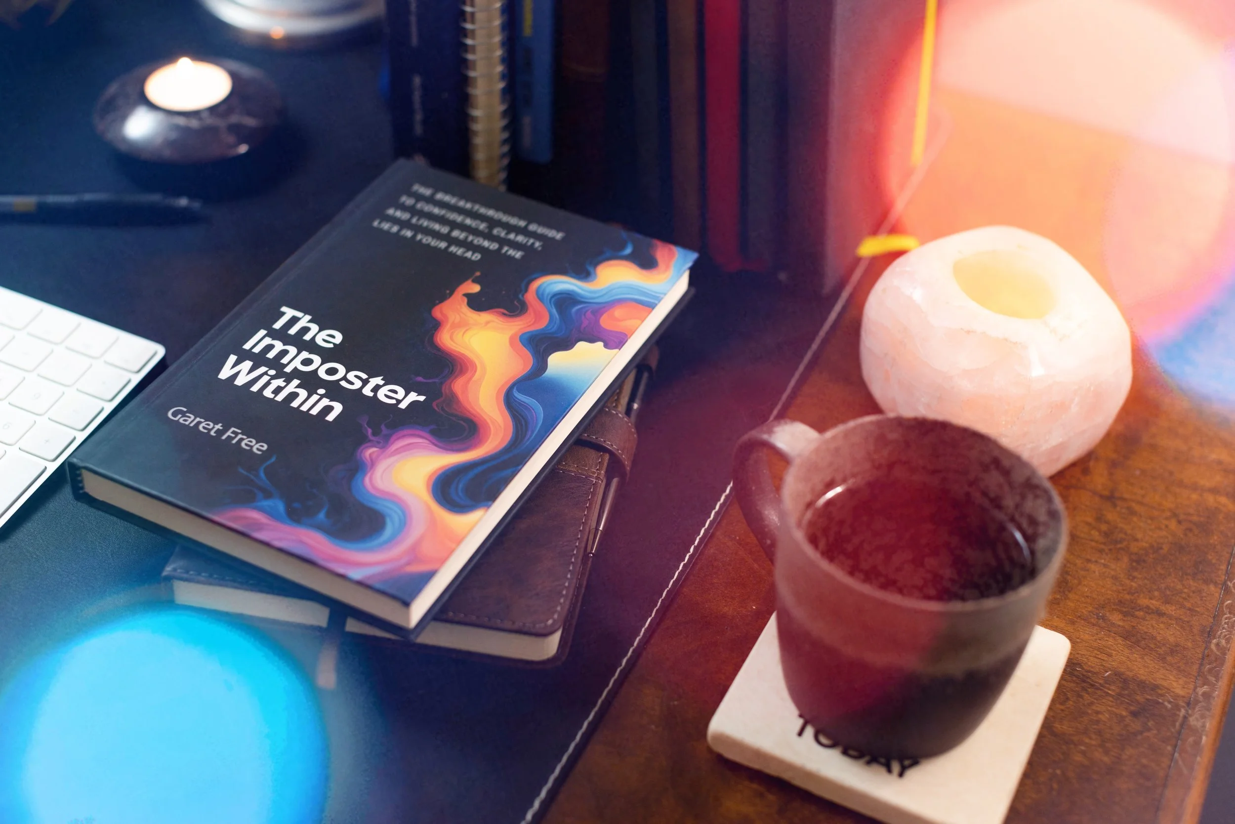 A desk scene with a book titled 'The Imposter Within' by Garet Free, a white keyboard, a pen, salt lamps, and a small cup in the foreground.