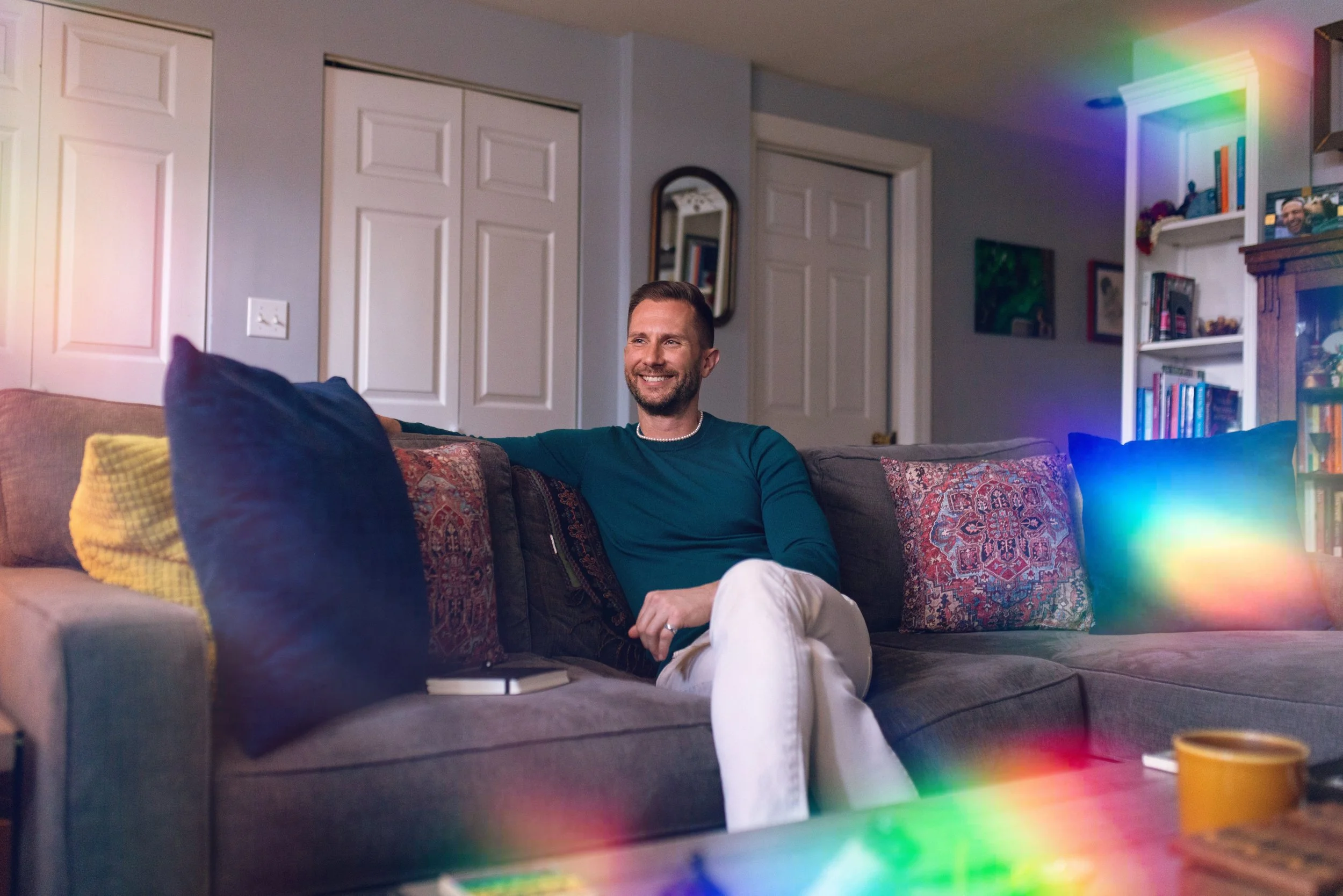 A man with a beard and short hair sitting on a gray couch with multicolored pillows, smiling, in a living room with white walls, a mirror, two closed closet doors, bookshelves, and rainbow-like light reflections.