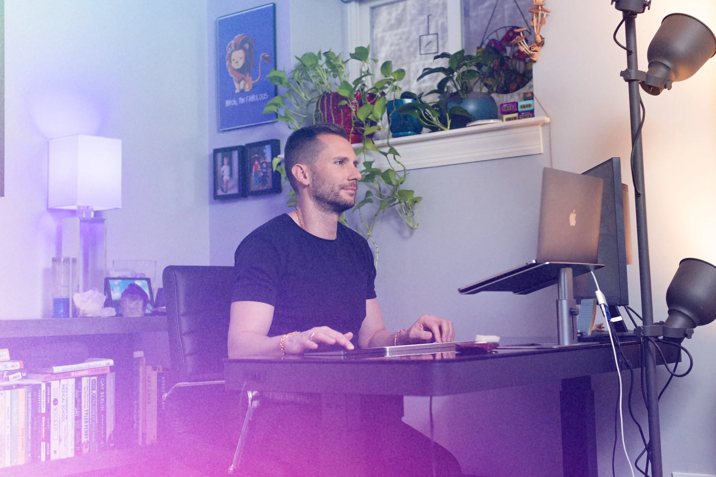 A man working at a desk in a room with modern decor. There are houseplants, framed pictures, books, a white lamp, and a professional studio light in the background.