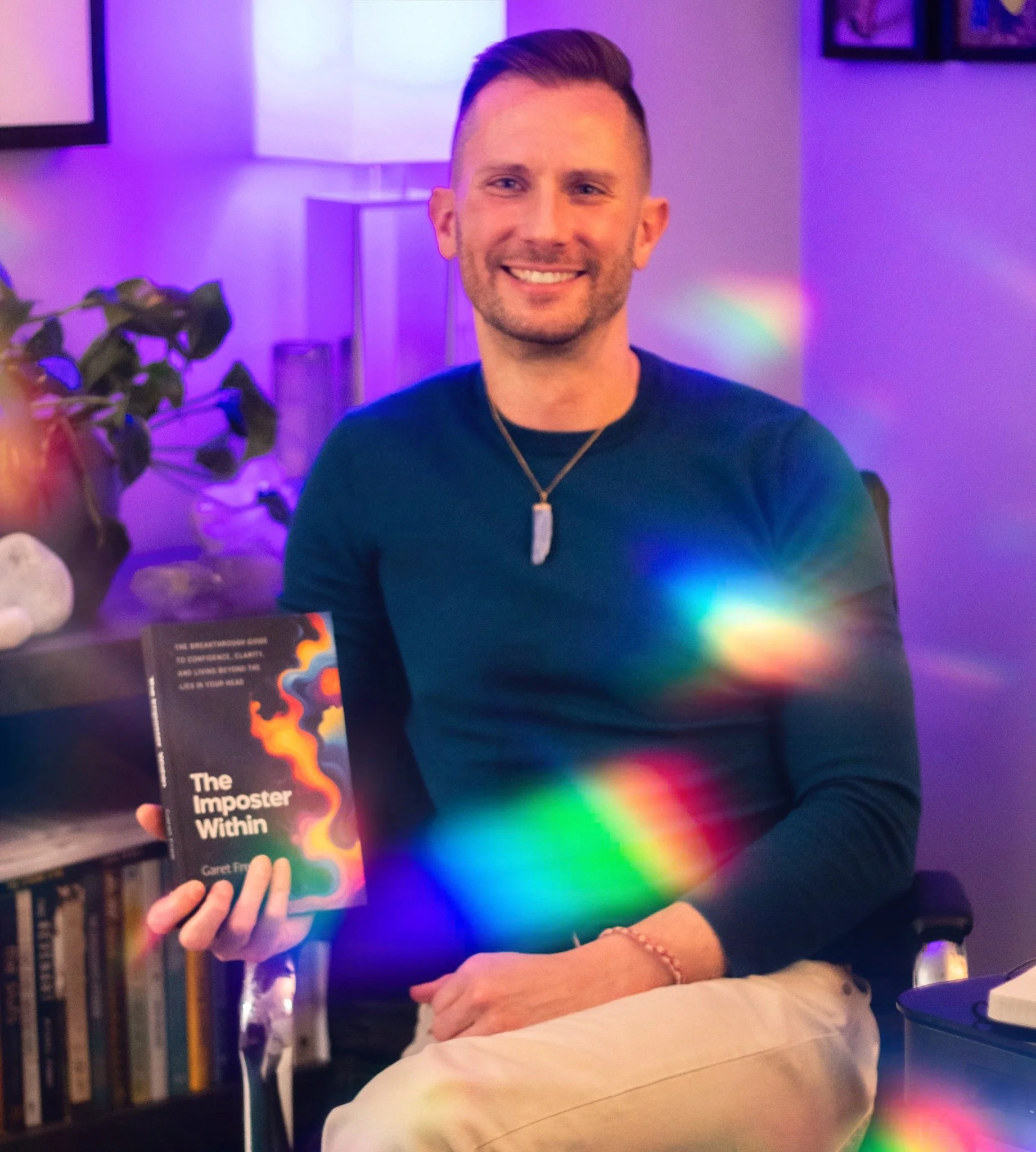 A smiling man holding a book titled 'The Imposter Within' in a room with purple lighting, a plant, bookshelves, and framed pictures on the wall.