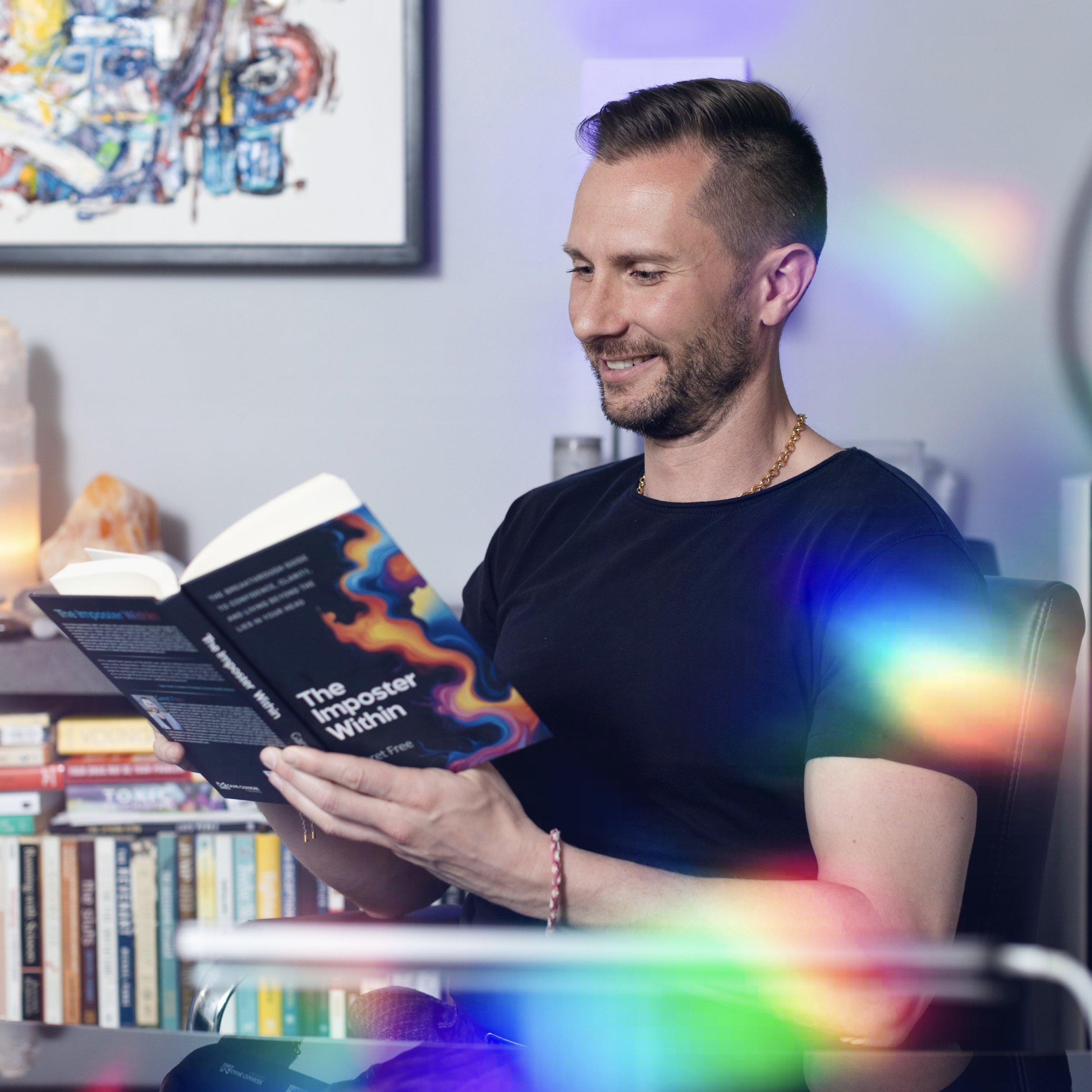 A man with short, dark hair and a beard is smiling while reading a book in a library or bookstore. He is wearing a black shirt and a gold chain necklace. In the background, there are shelves filled with colorful books and a framed abstract artwork on the wall. There is also a rainbow-colored light effect in the image.