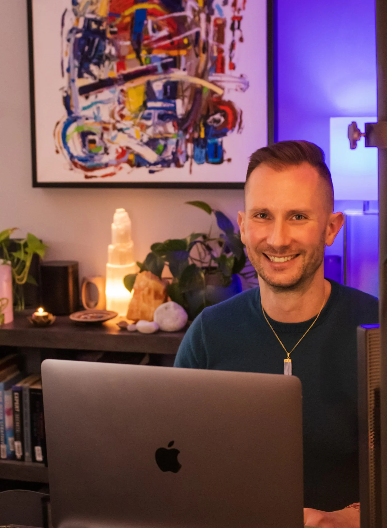 A man smiling sitting in front of a silver Apple iMac with a colorful abstract painting on the wall behind him. The room is dimly lit with blue and purple lighting, a small table with plants and candles visible in the background.