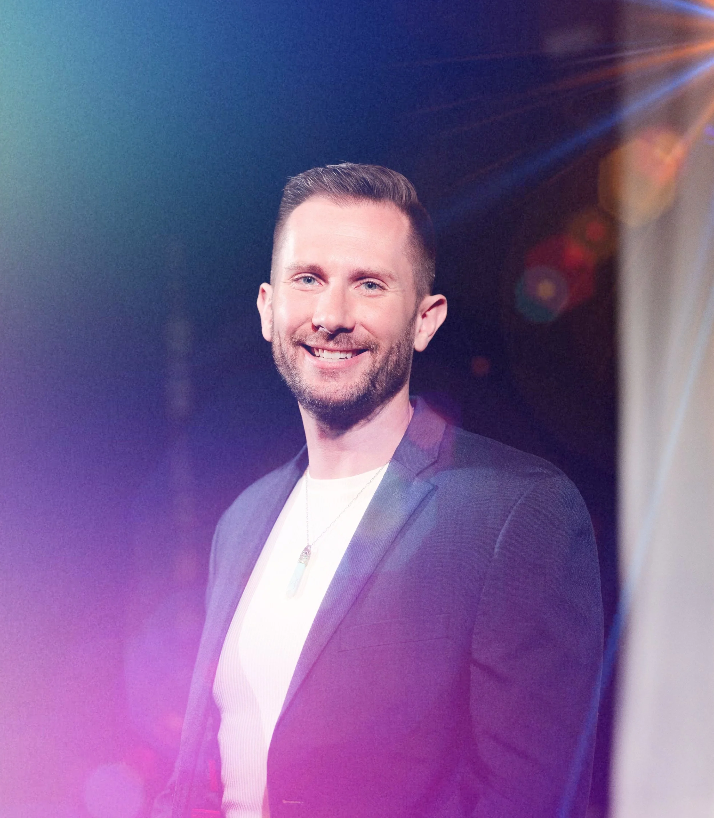 Partial view of a smiling man with short hair, beard, wearing a dark blazer, white shirt, and necklace, against a colorful background with lens flare effects.