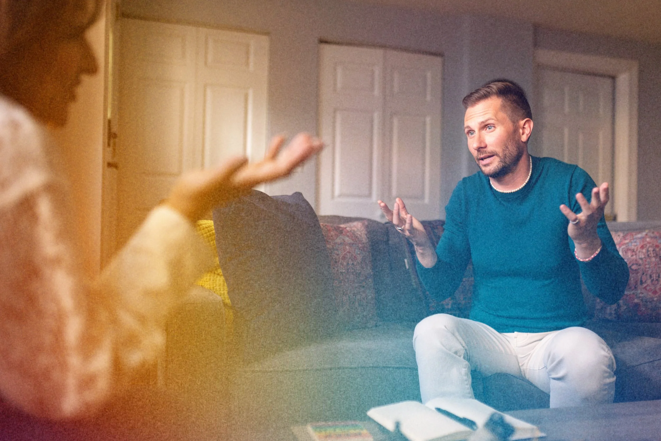 A man with short brown hair and blue eyes is sitting on a couch indoors, gesturing with his hands while talking. A woman with red hair is sitting nearby, but mostly out of the frame. The room has a door with paneling and a patterned pillow on the couch.