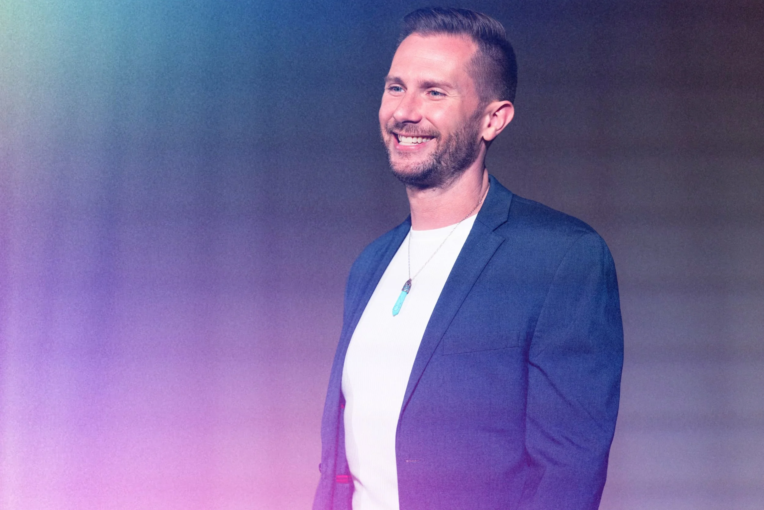 A man with short hair, a trimmed beard, and wearing a blue blazer and white shirt, smiling, against a gradient background.