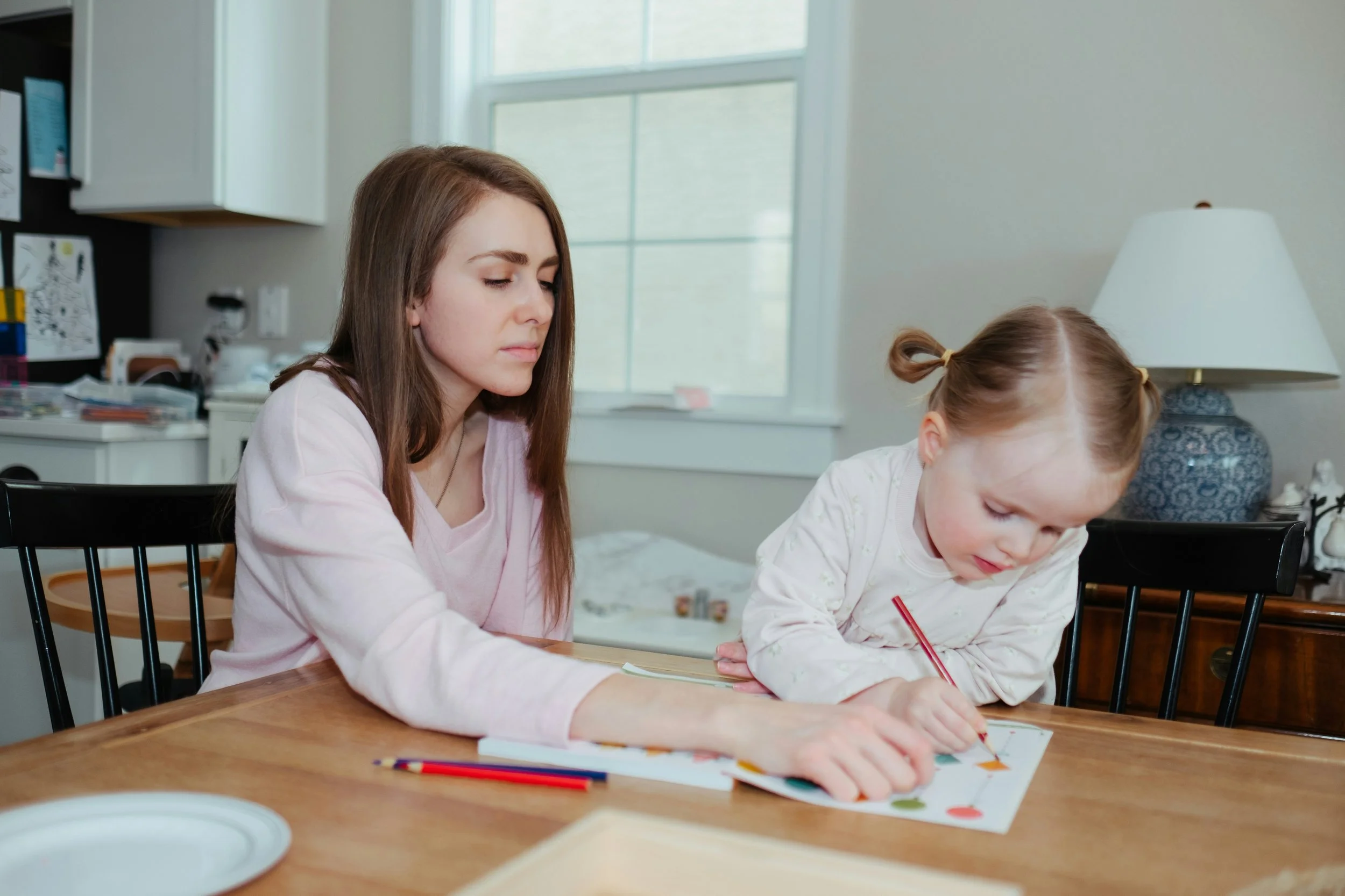 A young woman and a little girl sitting at a wooden table, drawing and coloring together in a bright room with a large window, a kitchen counter in the background, and a table lamp.