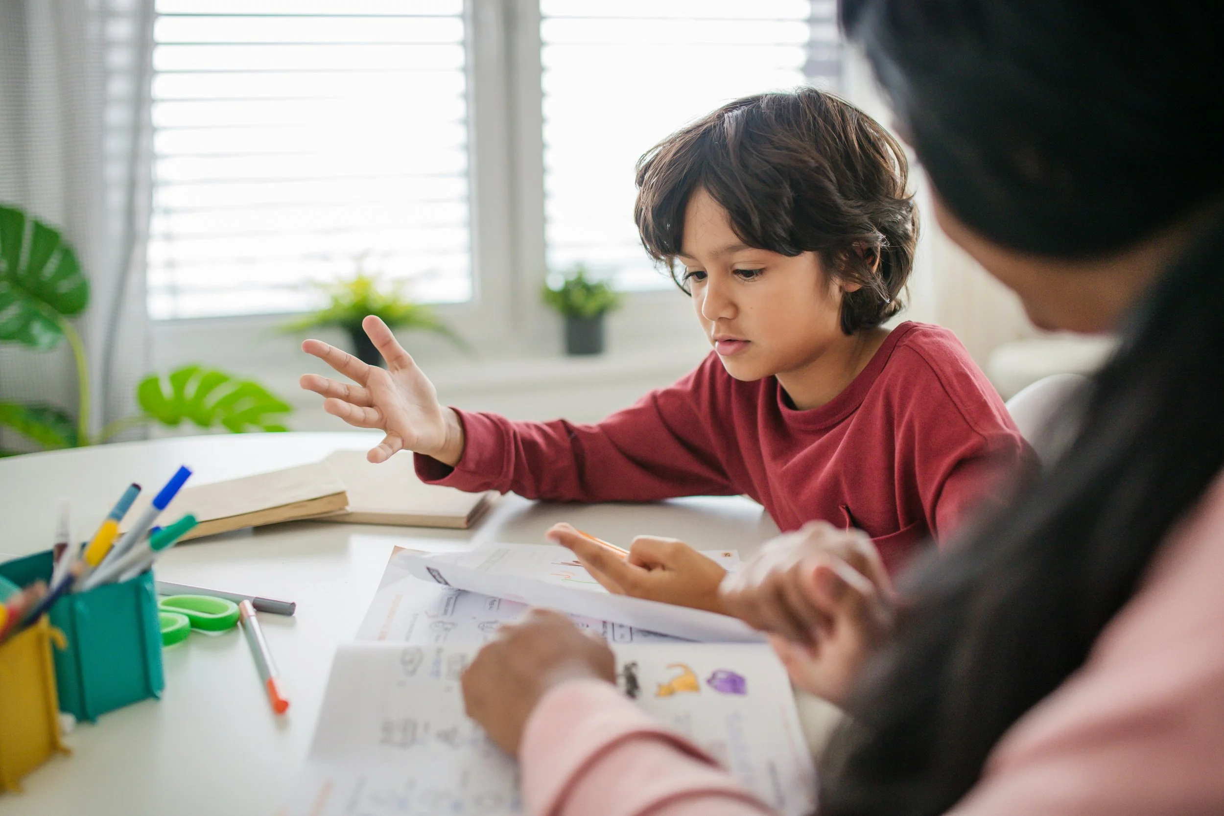 Child with dark hair wearing a red shirt sitting at a table, focusing on a worksheet, with another person partially visible. The table has colored markers, scissors, and papers. The background shows windows with blinds and some green plants.