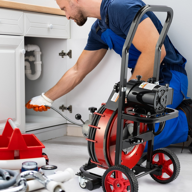 A plumber working under a kitchen sink, using a pipe inspection camera. The plumber is kneeling and wearing gloves, with plumbing tools and equipment nearby.