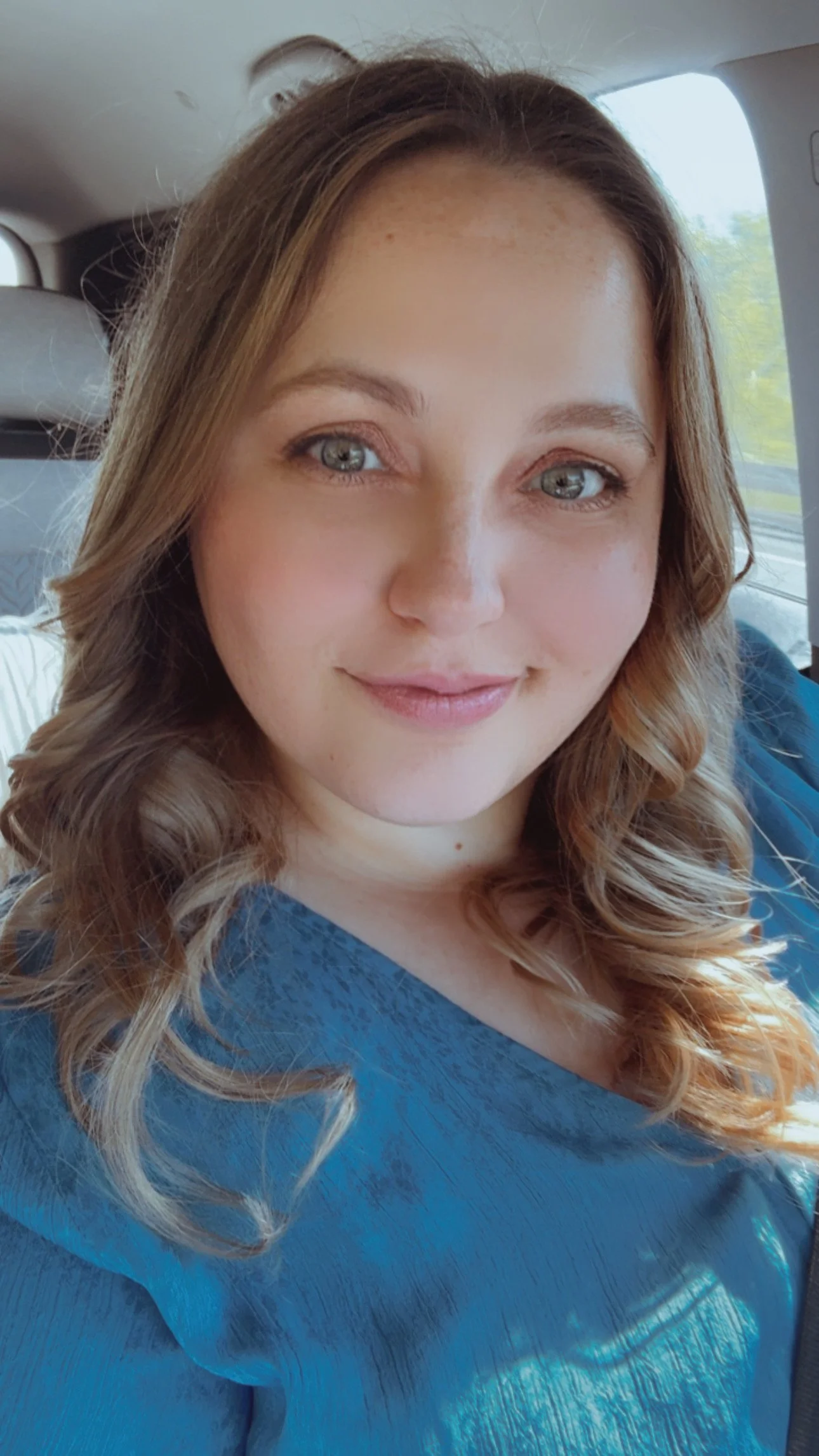 A woman with curly hair and blue eyes smiling while sitting in a vehicle, wearing a blue top.