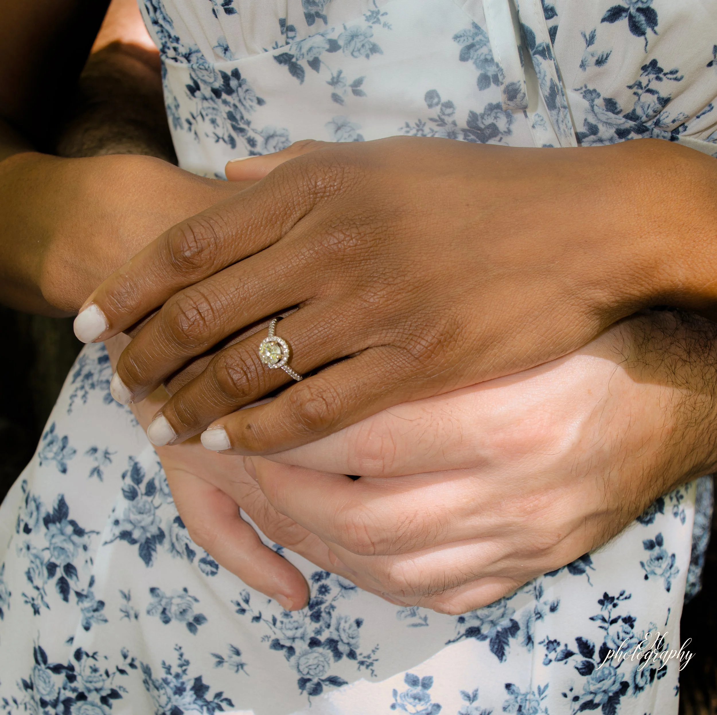 Close-up of two hands intertwined, one with a silver ring featuring a round diamond, resting on a blue floral patterned fabric.