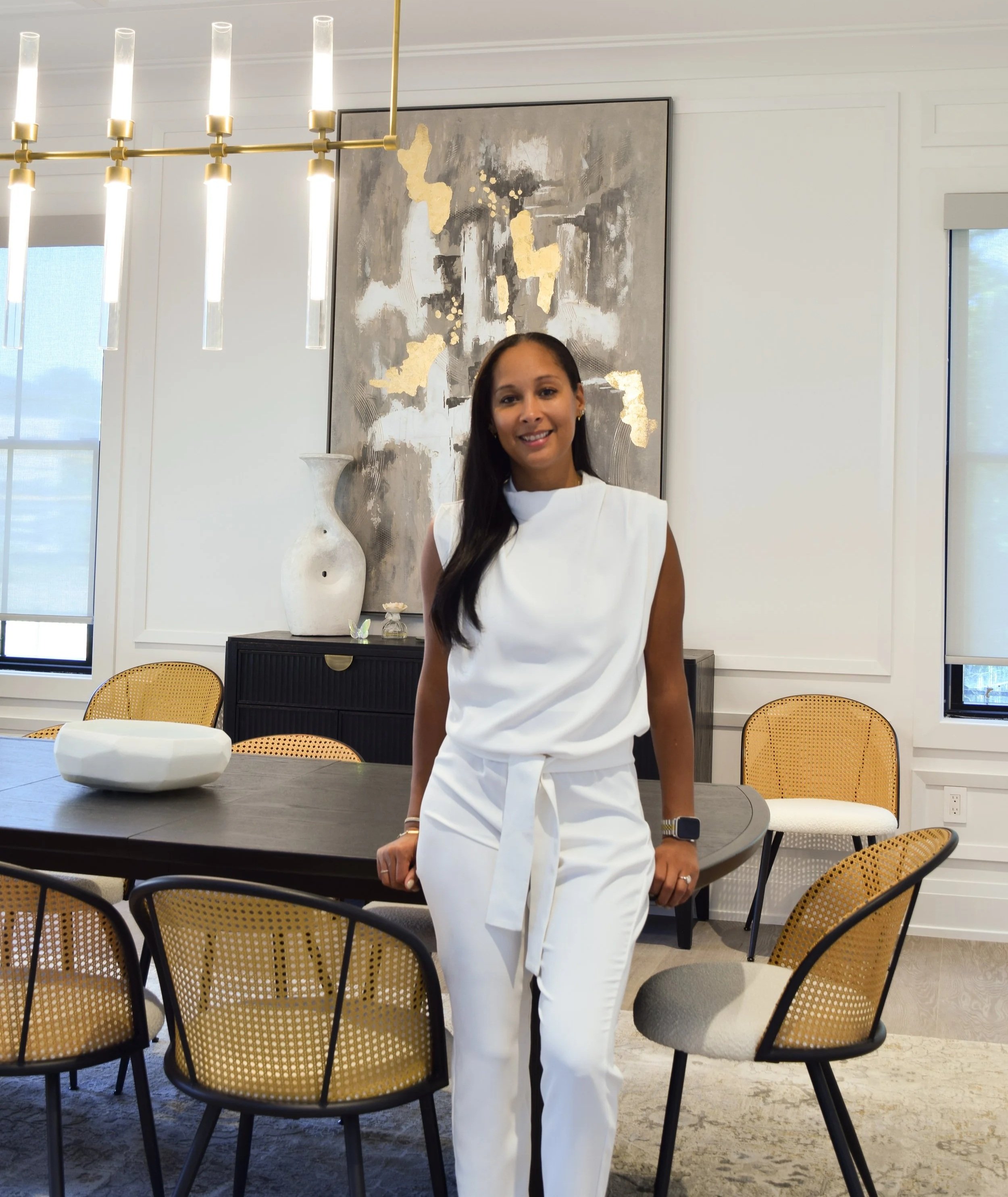 A woman with long dark hair standing in a modern dining room, smiling at the camera. She is wearing a white sleeveless top and white pants with a waist tie. The room has a large abstract painting with gold accents on the wall, a decorative white vase, and a black sideboard. There are several chairs with yellow woven backs and black frames around a dark dining table. A chandelier with cylindrical lights hangs from the ceiling, and large windows with shades are in the background.