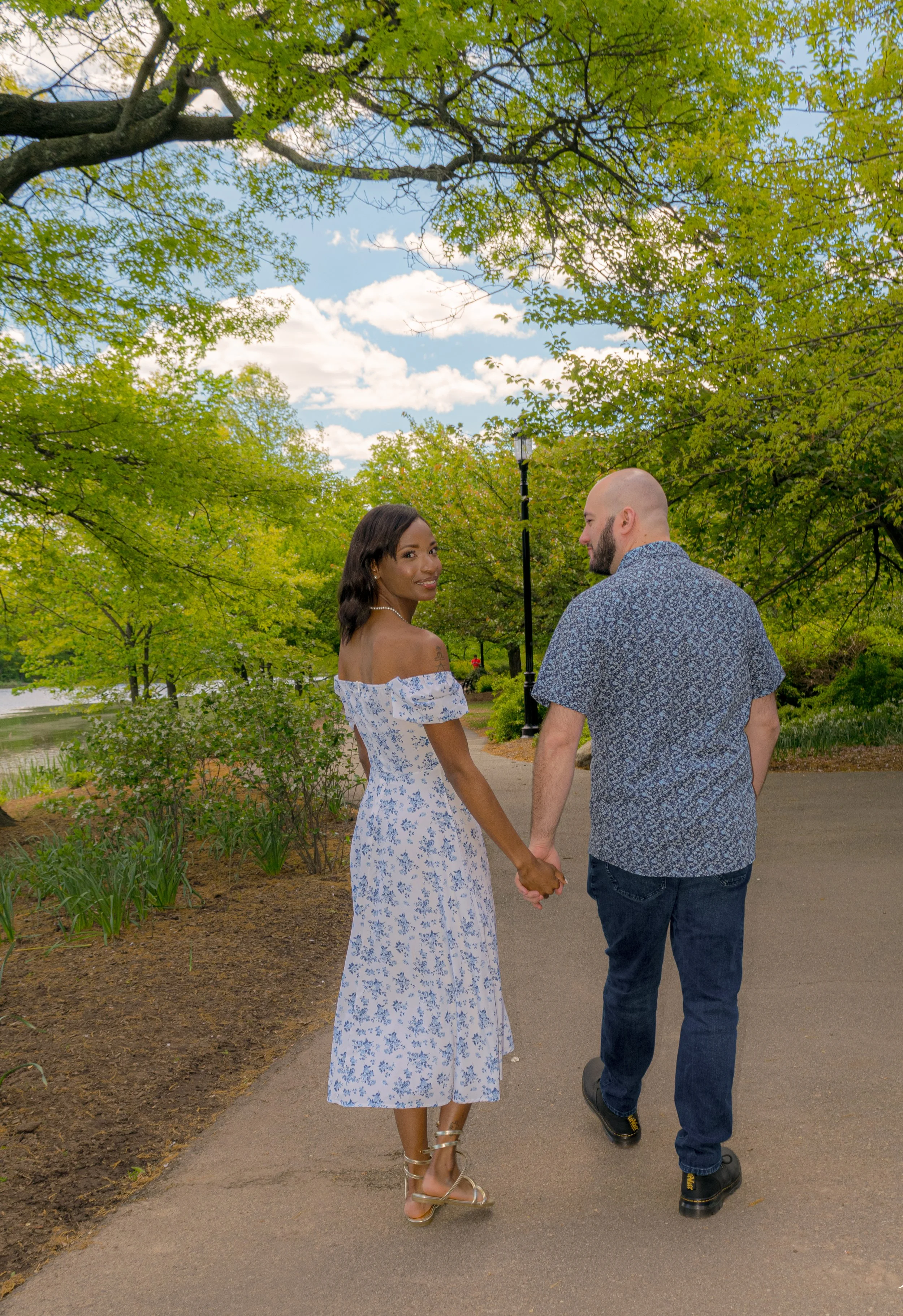 A couple holding hands and walking in a park surrounded by green trees and blue sky, with a woman smiling at the camera.