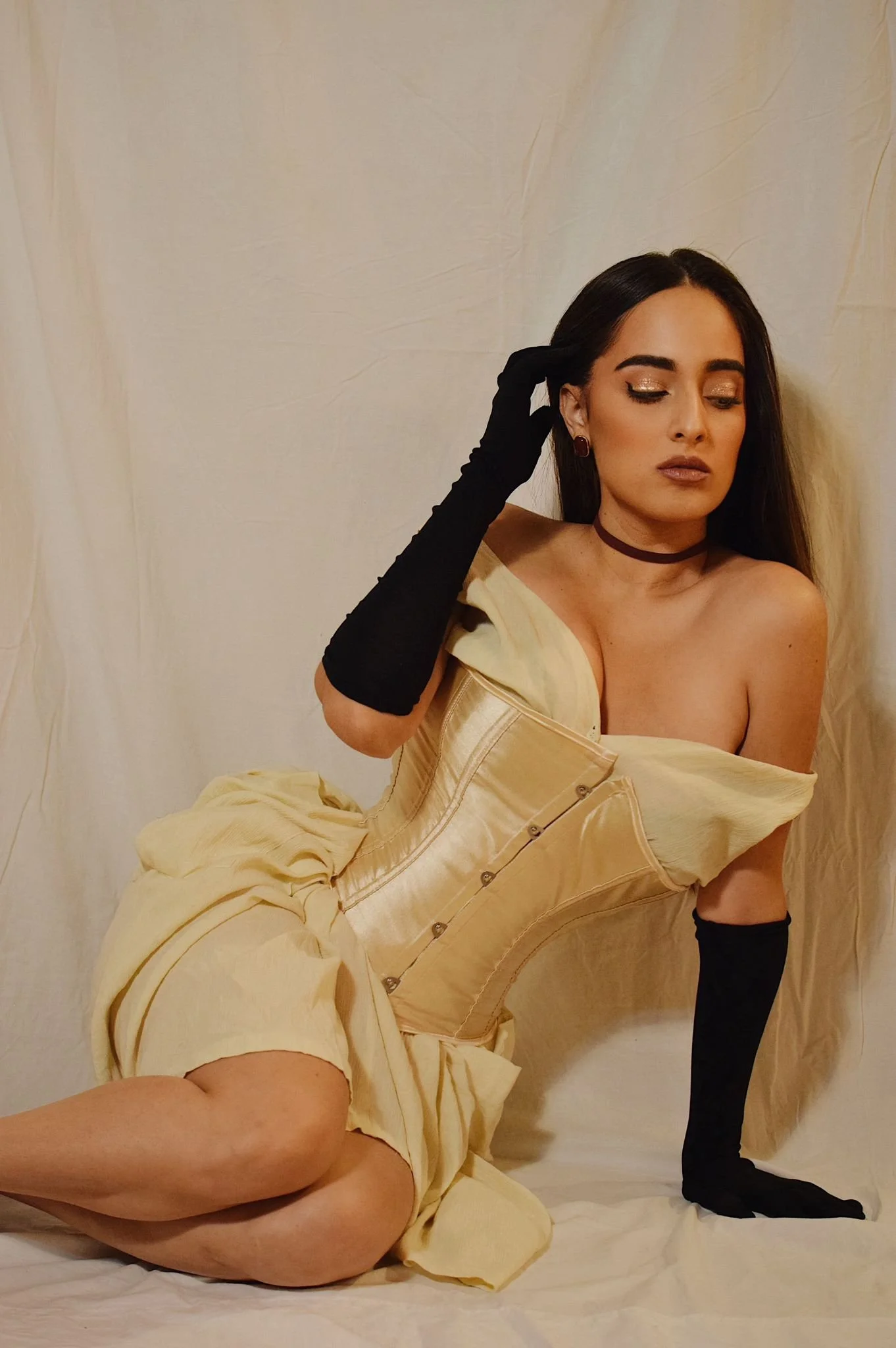 Mujer con vestido amarillo y guantes negros, posando con los ojos cerrados frente a una cortina beige.