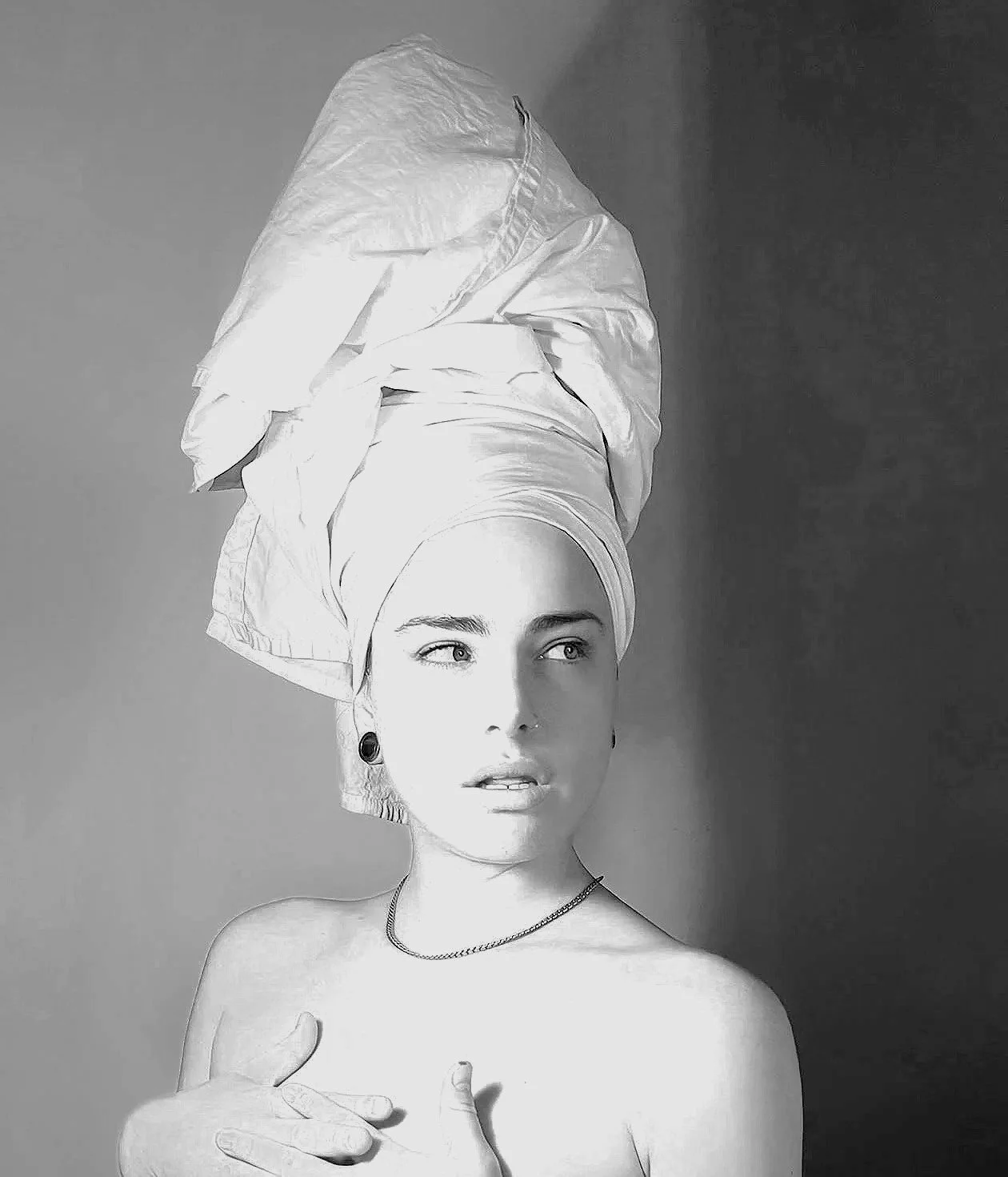 Mujer con tocado de tela enrollada en la cabeza, sin ropa, tocando su pecho y mirando hacia la izquierda, en una fotografía en blanco y negro.