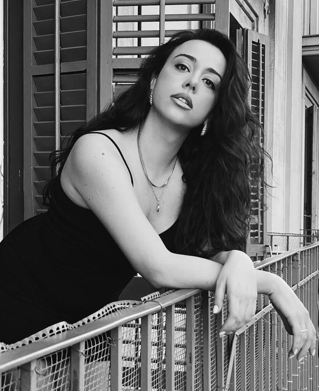 Retrato en blanco y negro de una mujer joven con cabello rizado, usando un vestido negro de tirantes y joyería, posando apoyada en una baranda en un entorno con lamas de madera en el fondo.