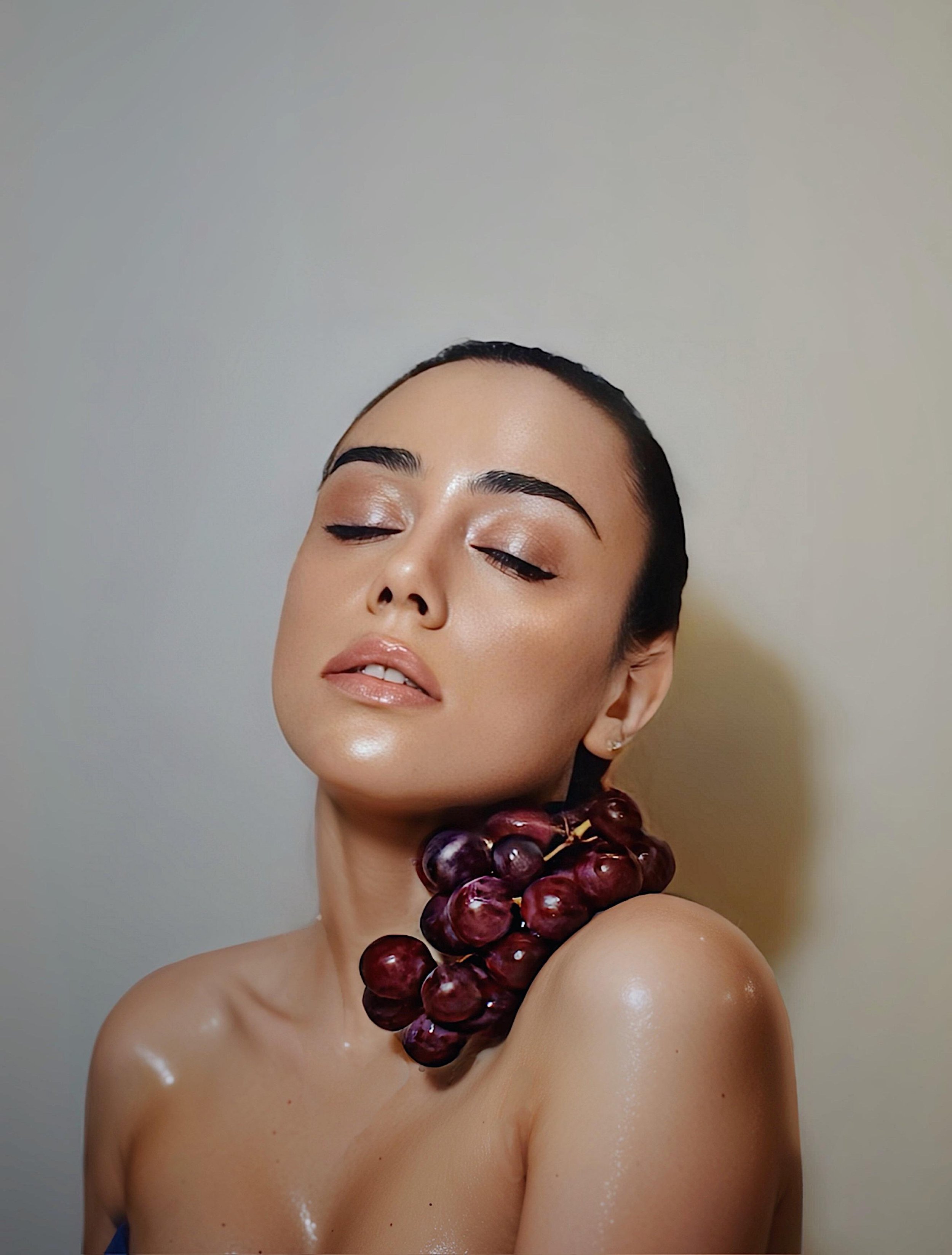 Retrato de una mujer con maquillaje y una corona de uvas en el cuello, posando con los ojos cerrados.