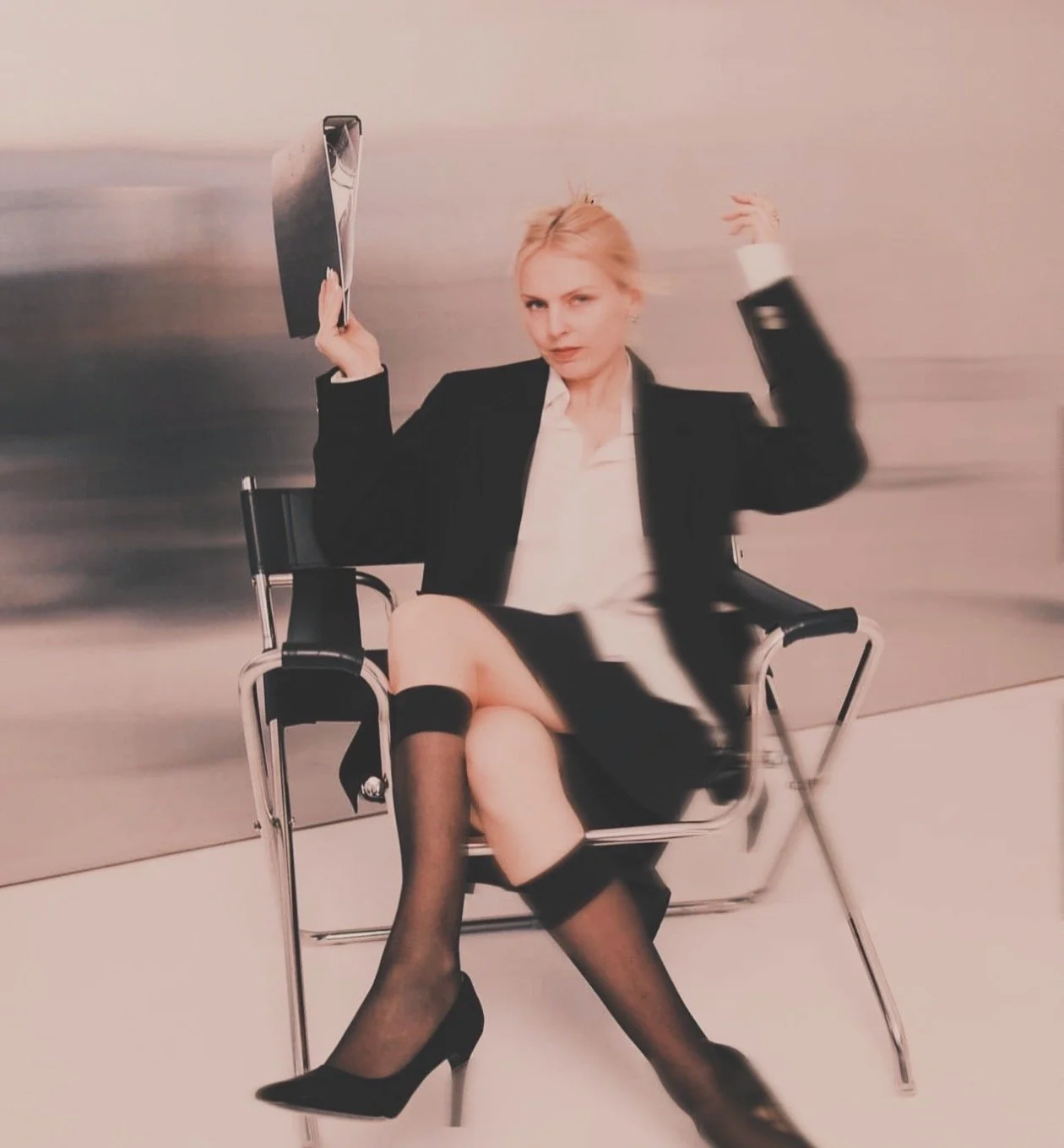Mujer sentada en una silla metálica, con uniforme de oficina, con medias de red y falda, sosteniendo un portafolio y ajustándose la media.