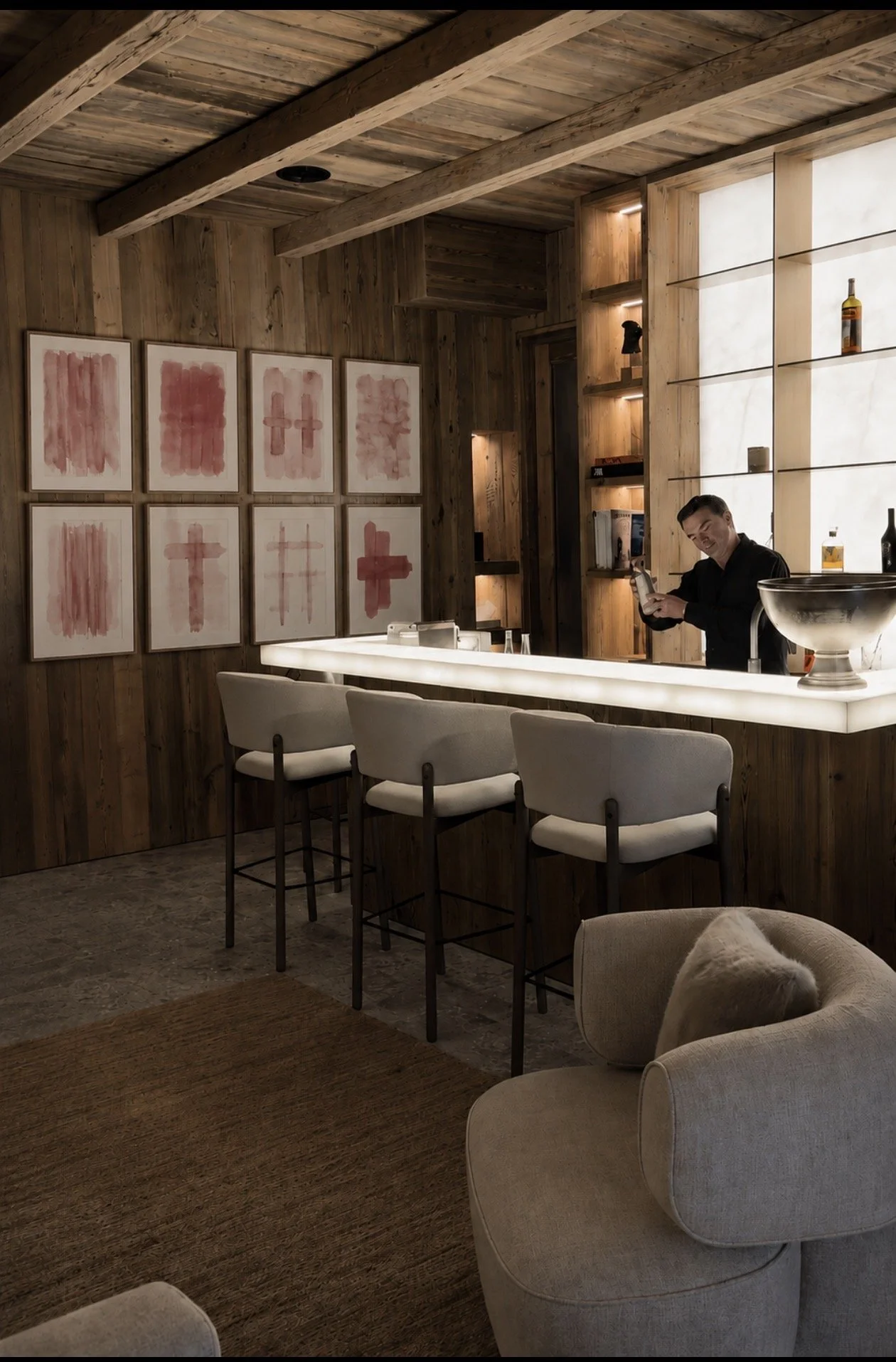 Un bar con paredes y techo de madera, decorado con cuadros abstractos en tonos rosados y rojos, una barra iluminada con cuatro sillas blancas, y una persona sirviendo una bebida.