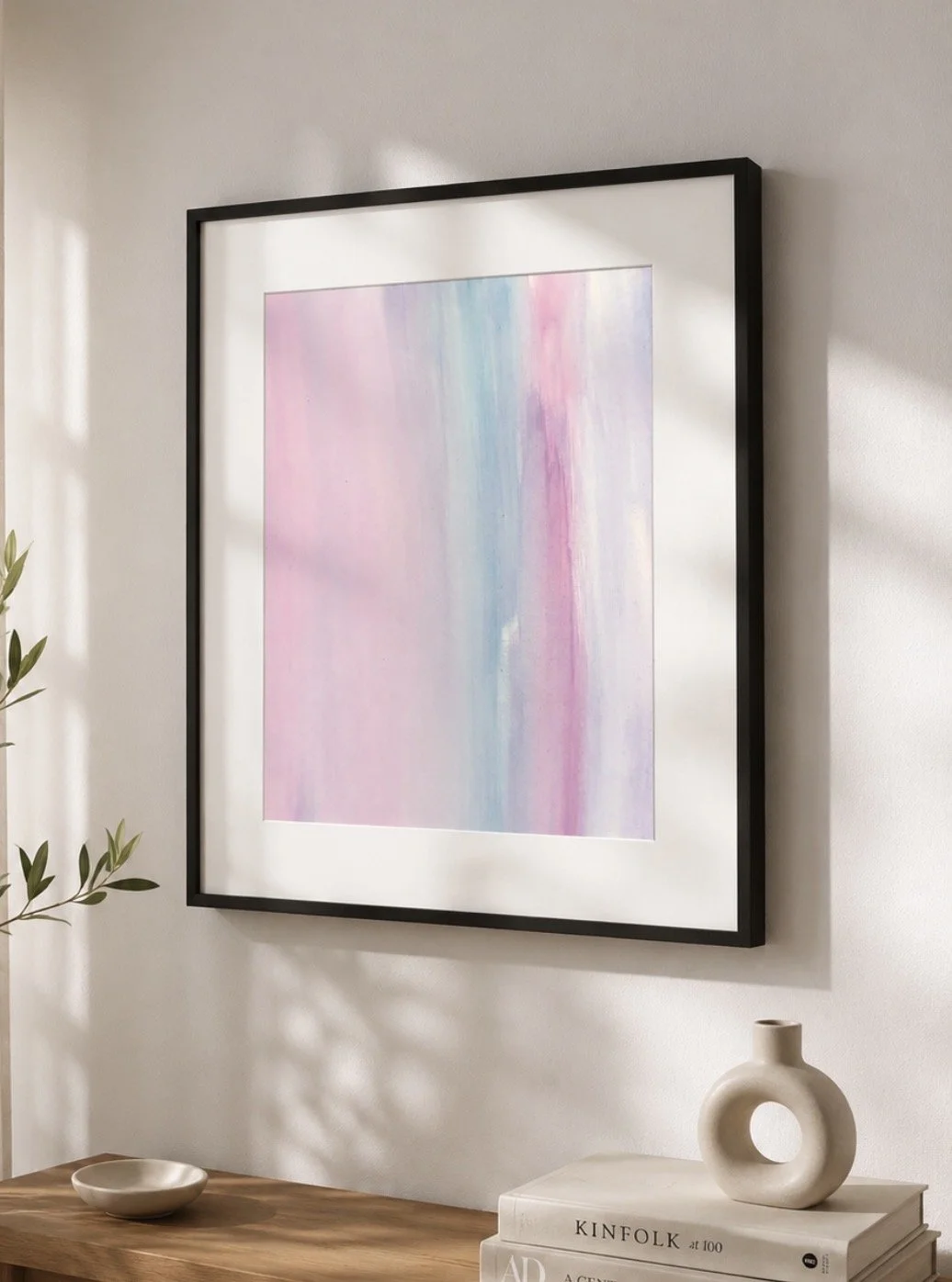 Cuadro abstracto con tonos pastel en un marco negro, colgado en una pared blanca, con una mesa de madera debajo que sostiene una vasija y unos libros.