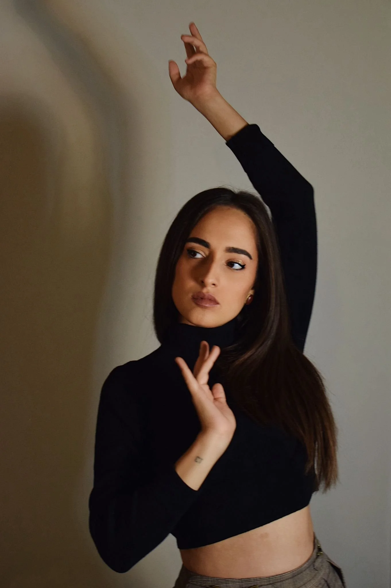 Retrato de una mujer joven con cabello largo y oscuro, usando un suéter negro con un escote distinto, levantando una mano y tocando suavemente la pared, con expresión introspectiva.