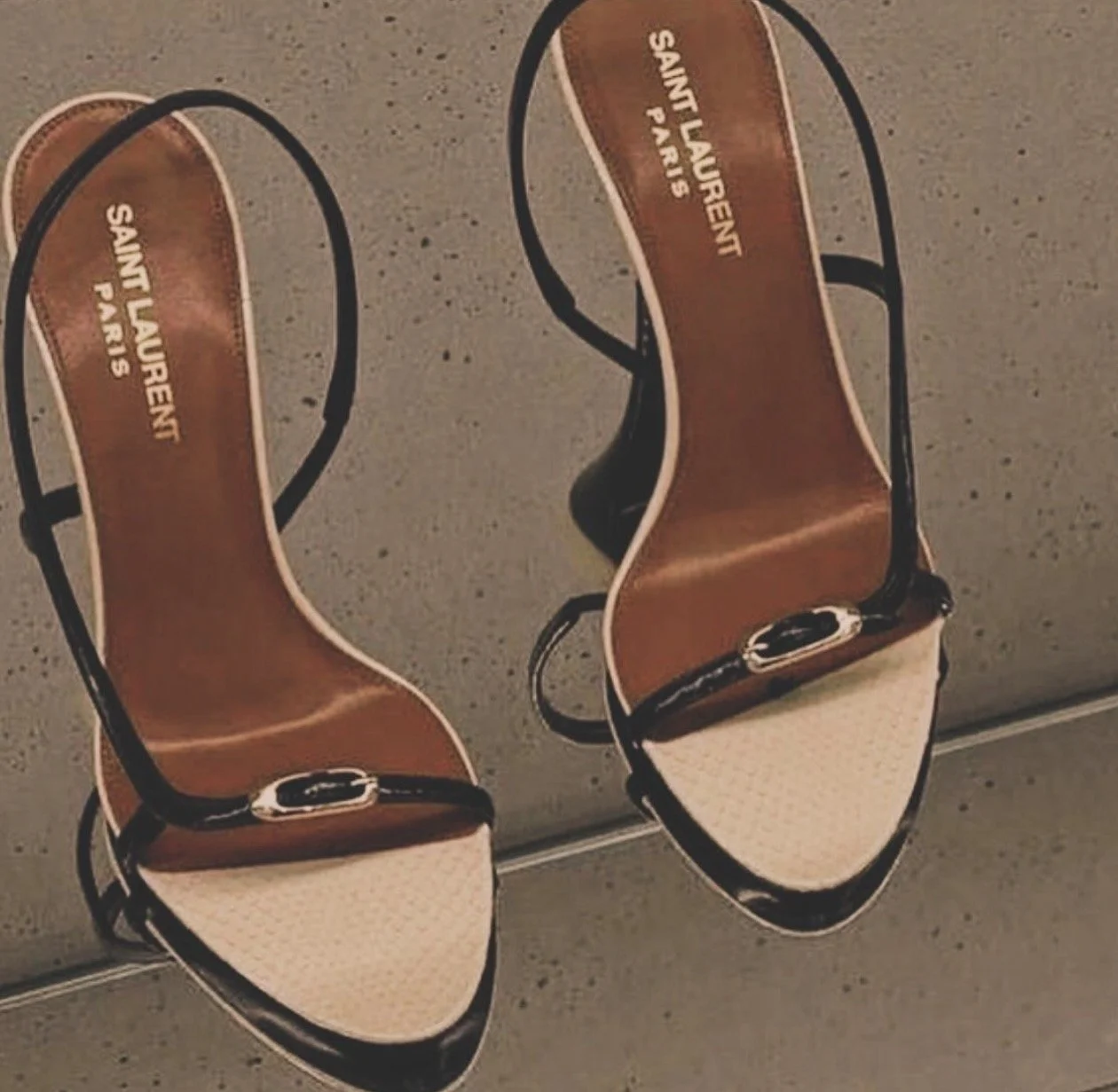 Sandalias de tacón con diseño de tiras negras y punta en tono beige, marca Saint Laurent Paris, colocadas sobre un suelo de color gris.