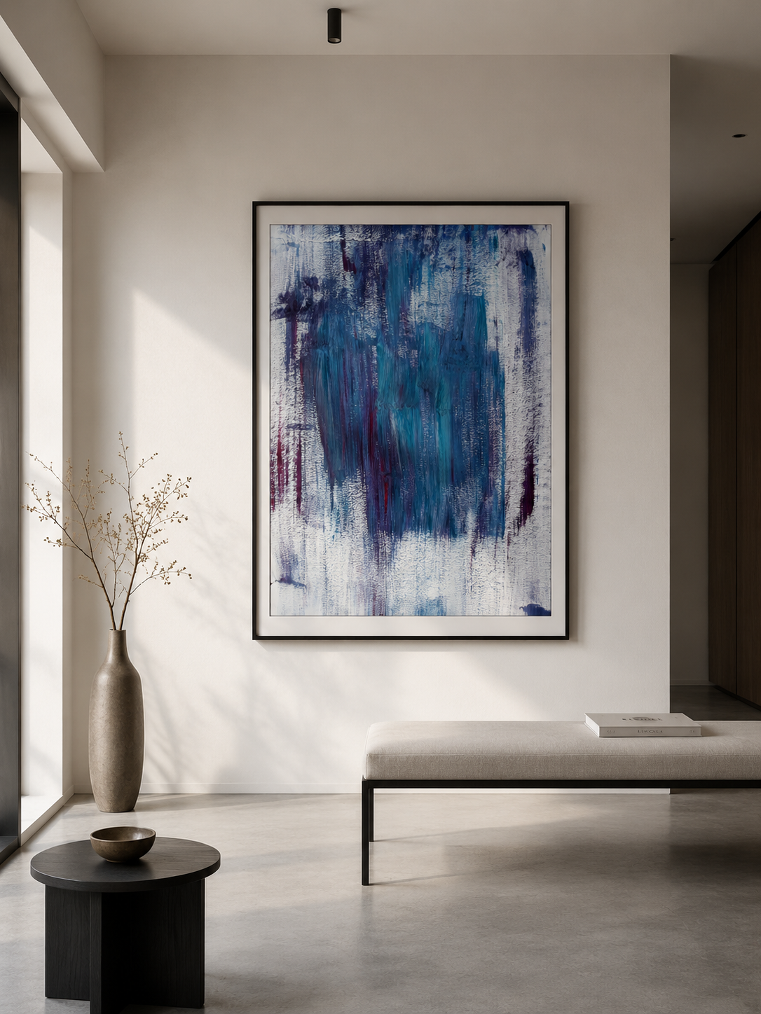 Interior moderno con una pintura abstracta en tonos azules y morados, un banco con cojín beige, una consola negra con libros y un jarrón con ramas secas, todo en un espacio con paredes blancas y luz natural.