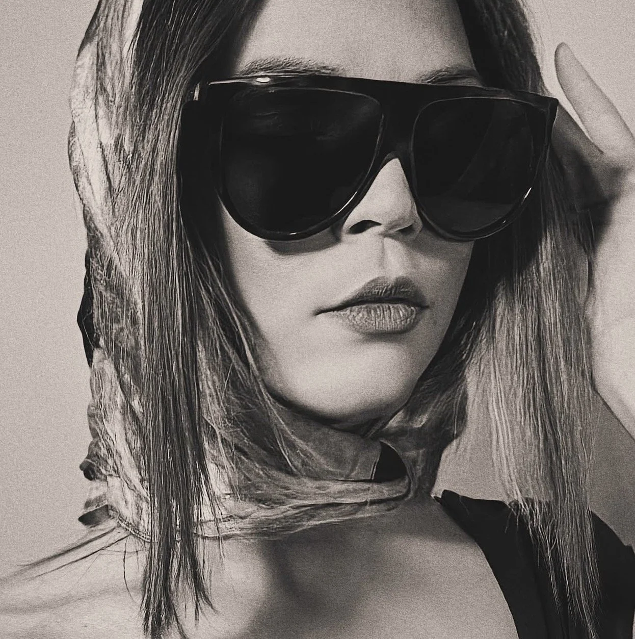 Retrato en blanco y negro de una mujer con cabello suelto, usando gafas de sol grandes y una bufanda en el cuello.