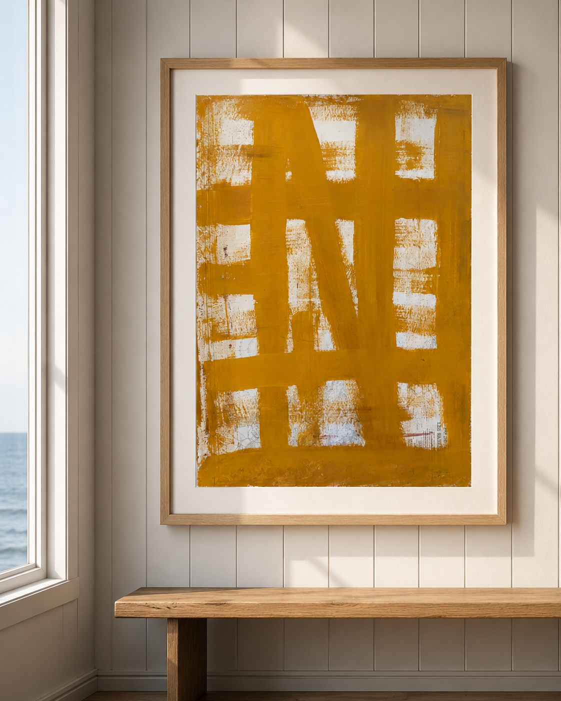 Cuadro decorativo con letras de color amarillo mostaza en un marco de madera, colgado en una pared blanca panelada, con una ventana a la izquierda y vista al mar en el fondo.