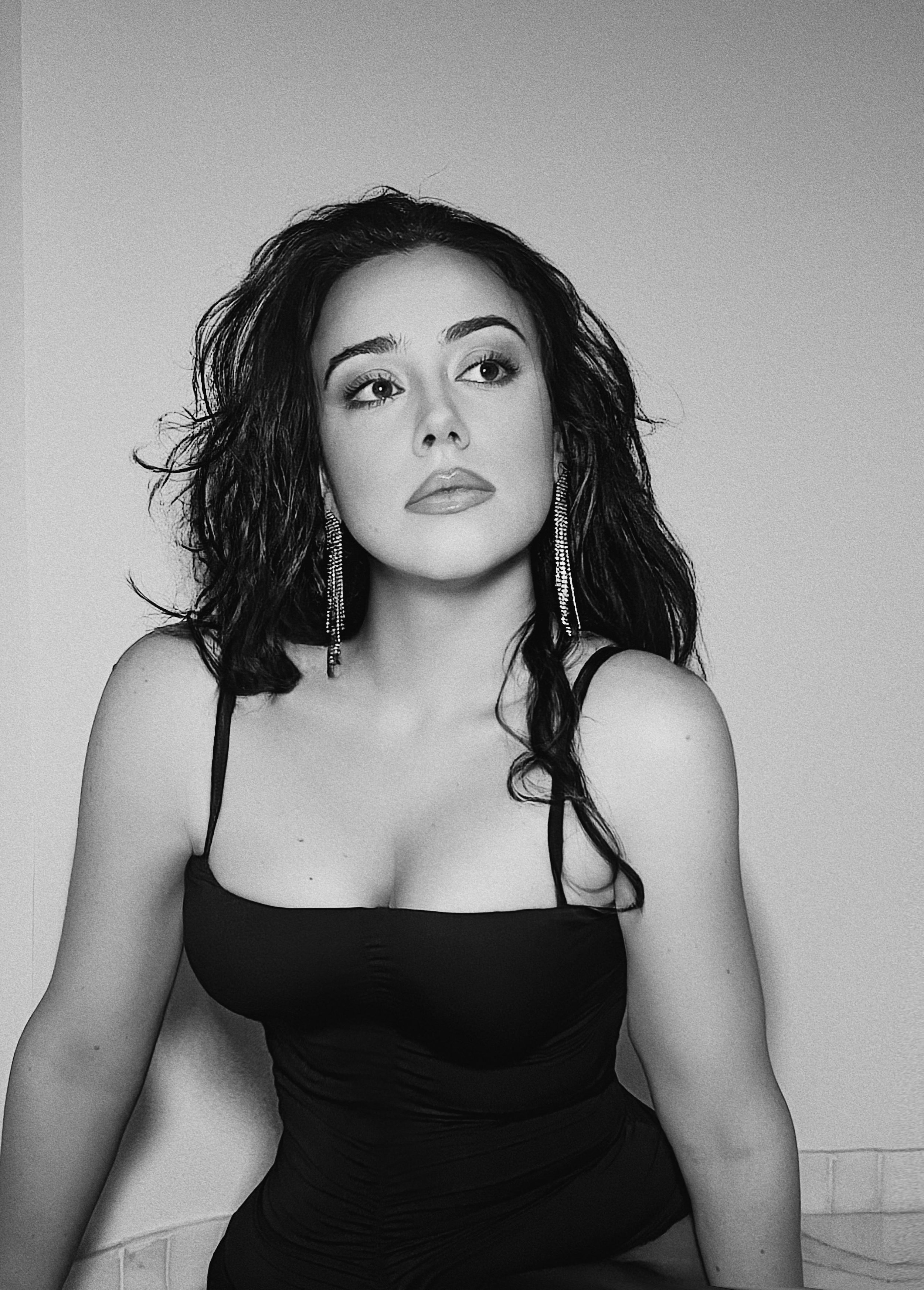 Retrato en blanco y negro de una mujer con cabello rizado y largo, usando un vestido ajustado, con pendientes largos, posando en un ambiente interior. 