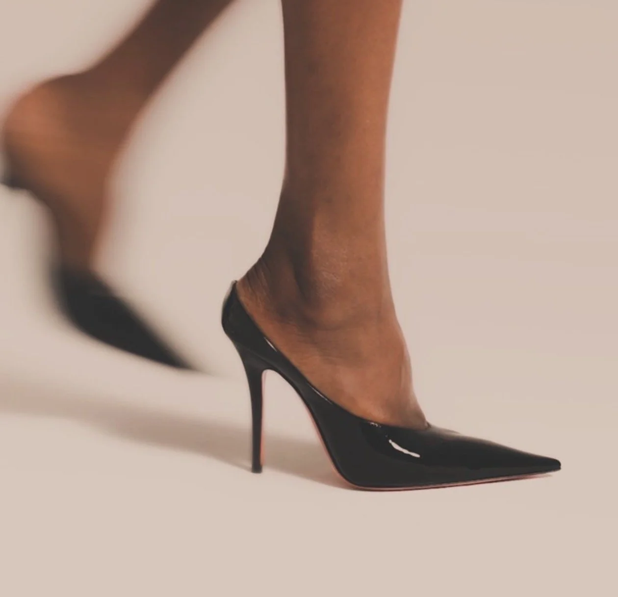 Pierna femenina con zapato de tacón negro y punta puntiaguda sobre fondo claro.
