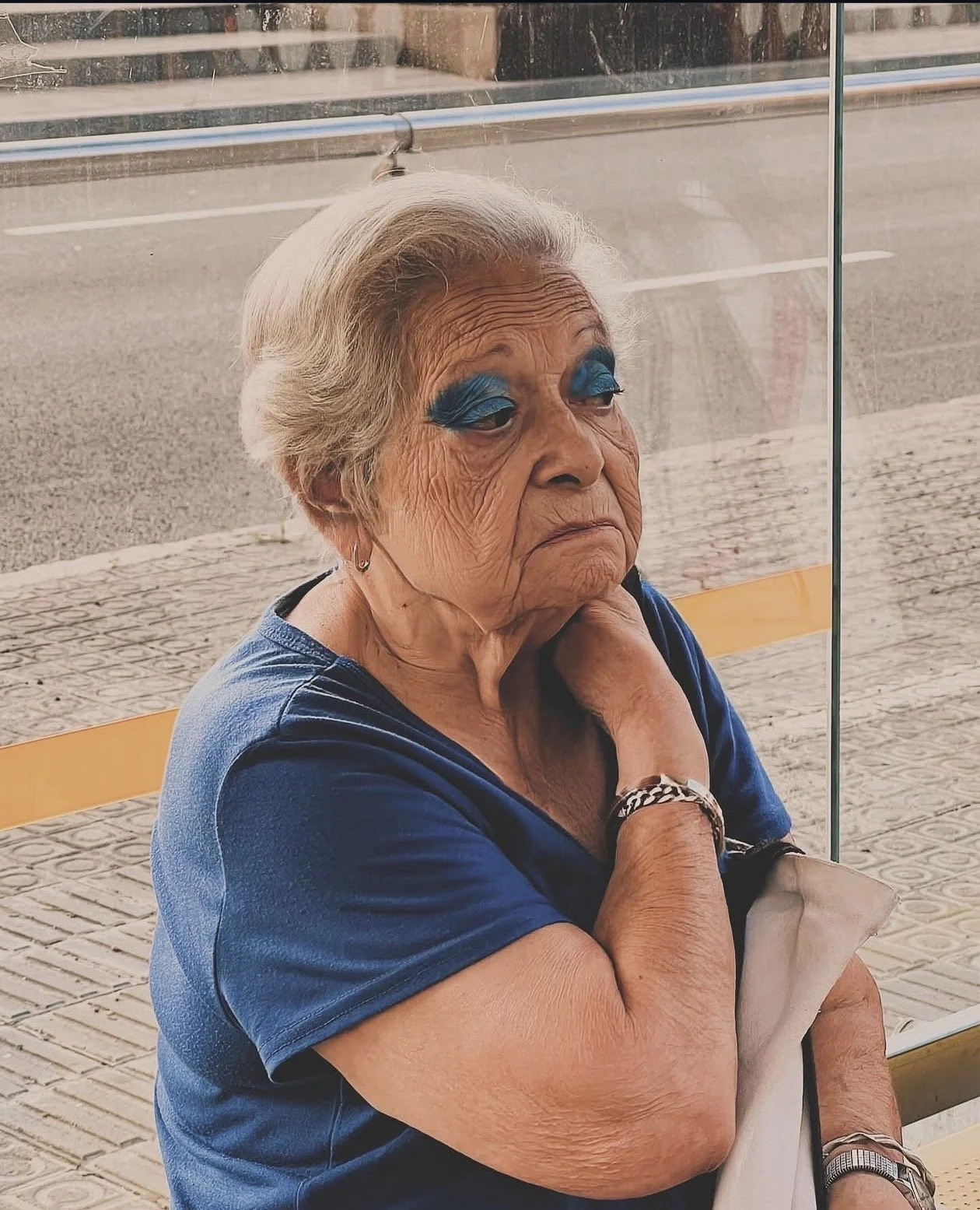 Anciana de cabello canoso, vistiendo camiseta azul, con maquillaje llamativo en los ojos, sentada en una banca con fondo de acera y vidrio.