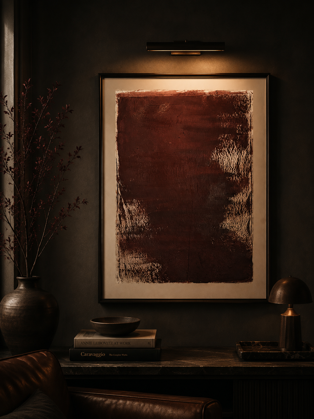 Una pared oscura con un cuadro abstracto en tonos marrones y negros, iluminado por una luz encima. A la izquierda, una jarra con ramas y a la derecha, una lámpara de escritorio y algunos libros en una mesa.
