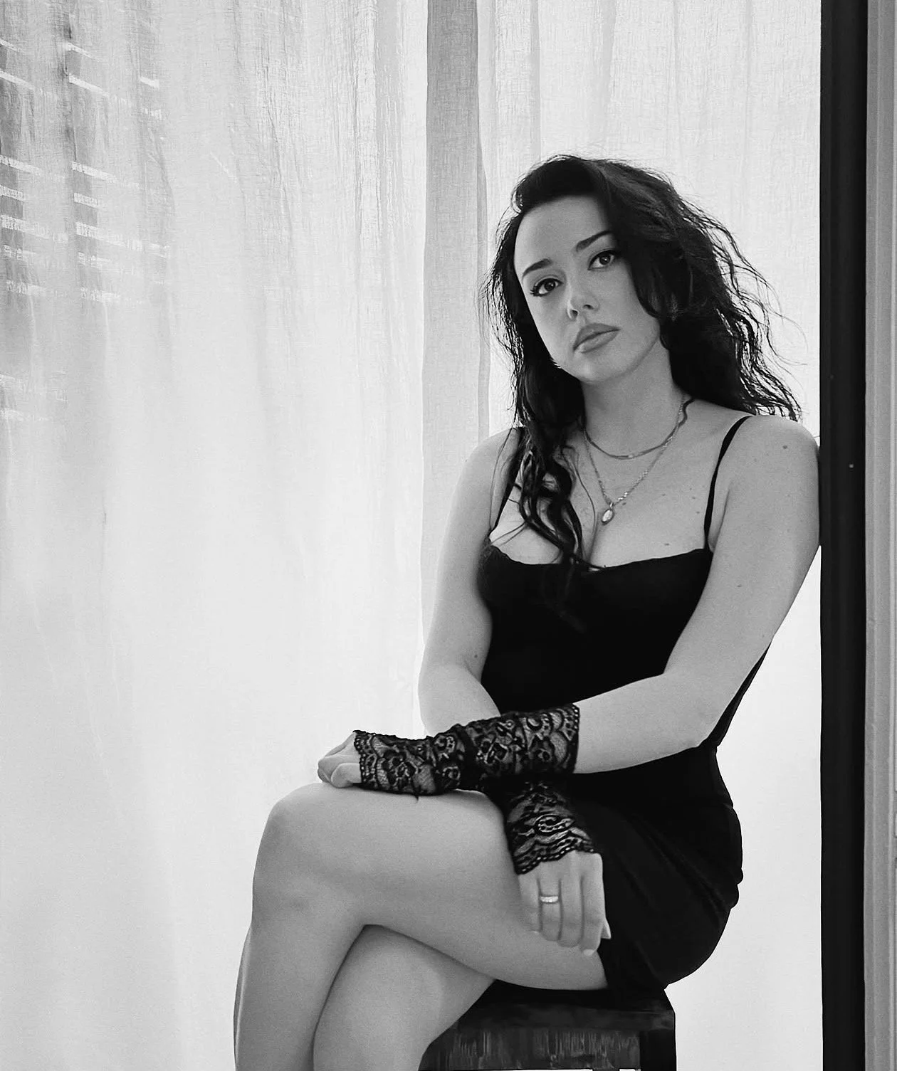 Retrato en blanco y negro de una mujer con vestido negro y guantes de encaje, sentada en un mueble junto a una ventana con cortinas, posando con expresión seria.