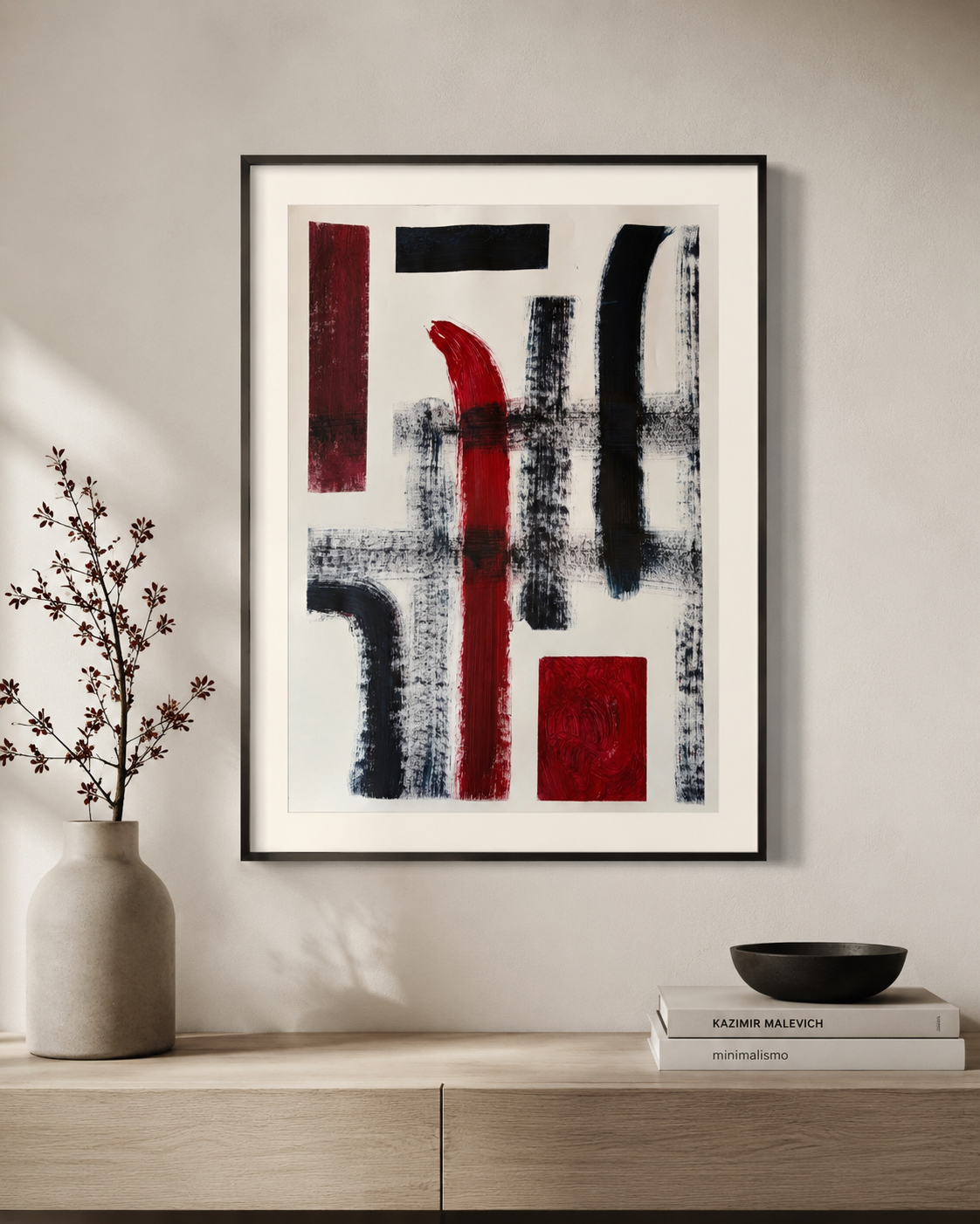 Cuadro abstracto con formas negras y rojas en una pared blanca, junto a una mesa con un jarrón y ramas secas, y libros y un recipiente negro.