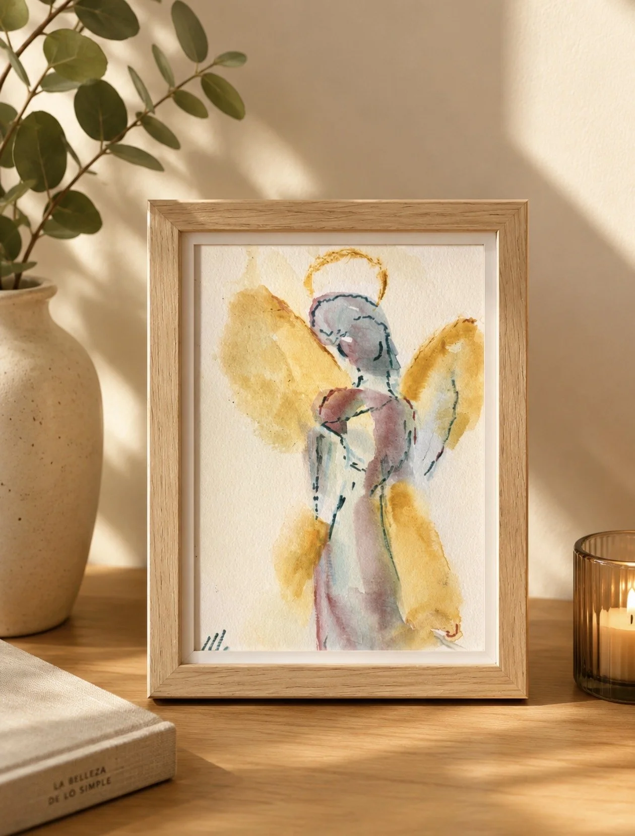Cuadro con pintura abstracta de un ángel con alas doradas, fondo beige, en un marco de madera. A la izquierda hay una maceta con planta de hojas verdes, y a la derecha, una vela en un vaso de cristal oscuro.