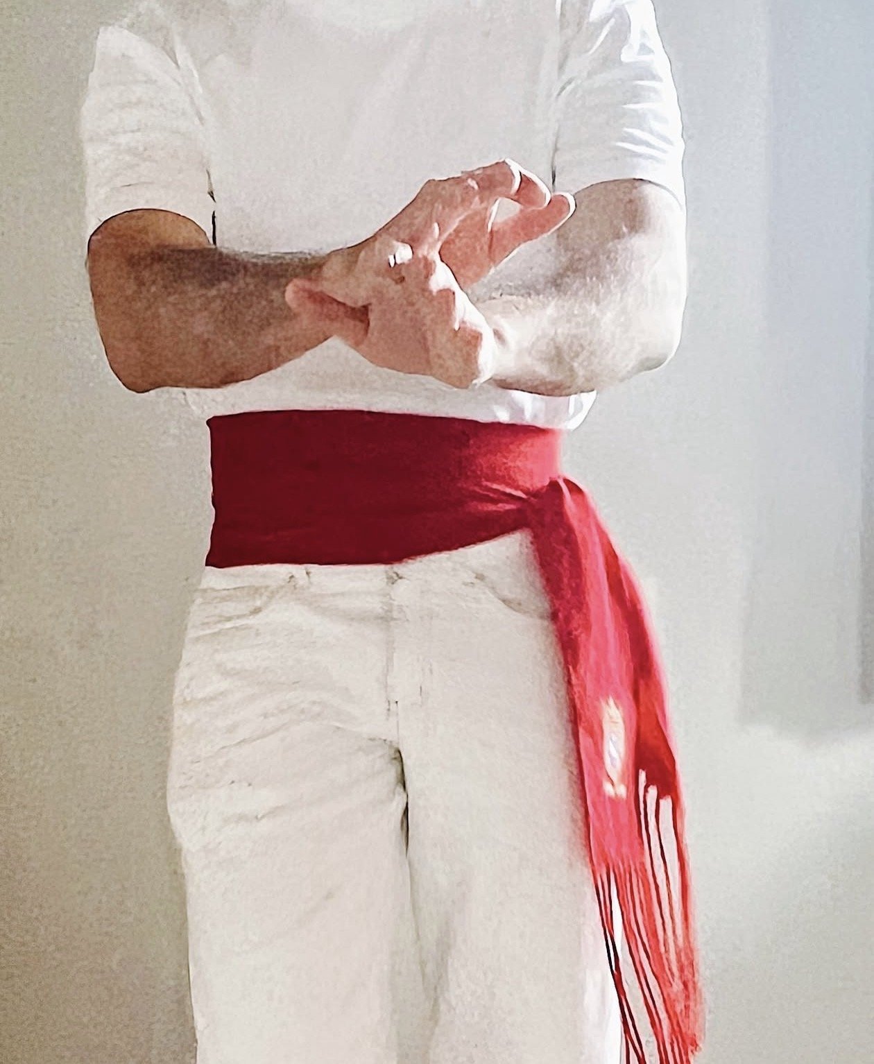 Persona vestido con camiseta blanca, pantalón blanco, y cinturón rojo atado con lazo y flecos.